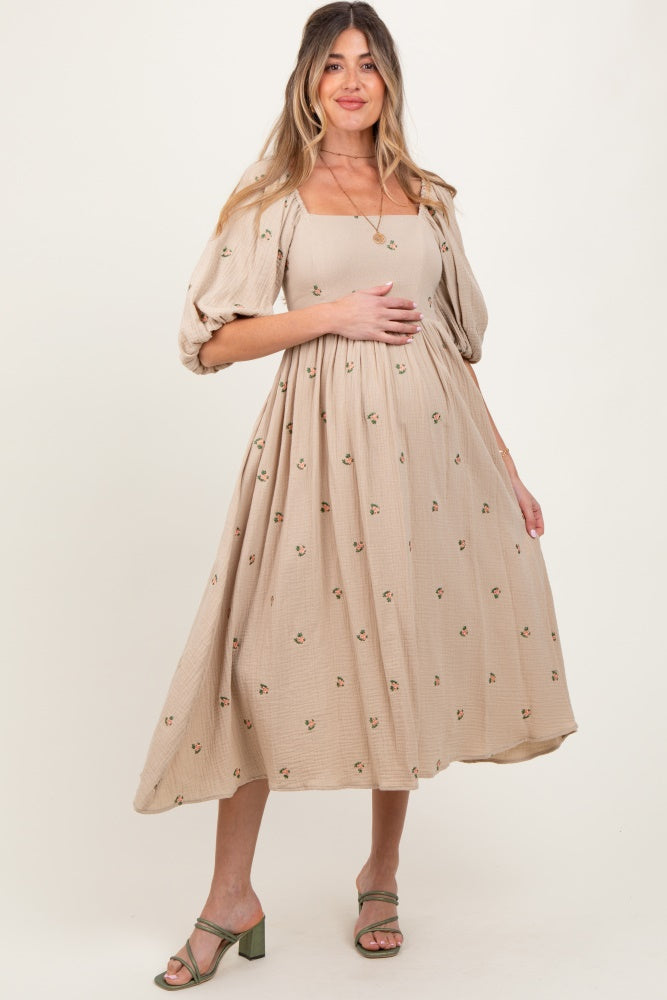 woman wearing a beige maternity dress with puff sleeves and floral embroidery, holding her baby bump