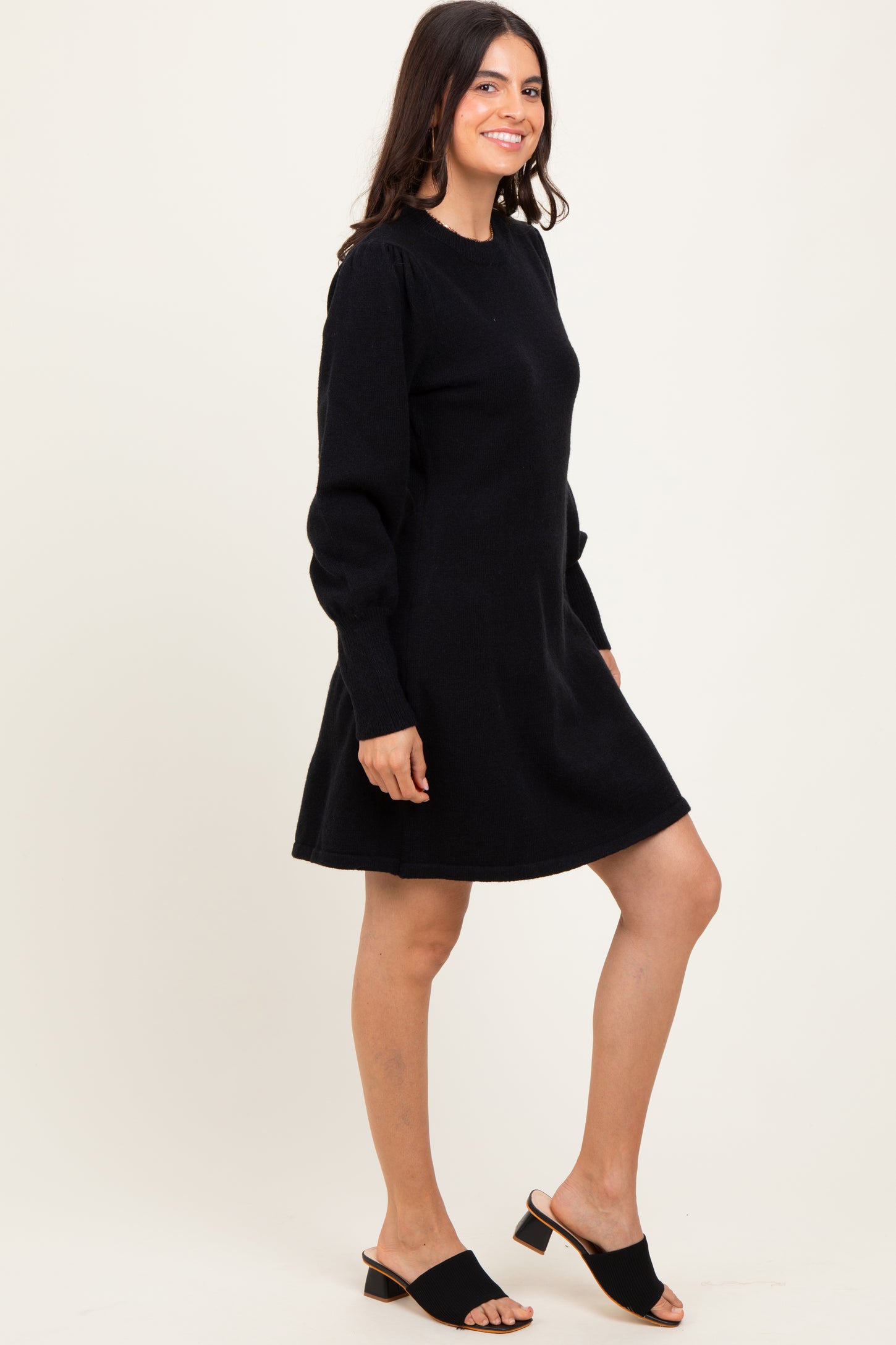woman wearing a black long sleeve sweater dress and black slide sandals, standing sideways with a relaxed posture