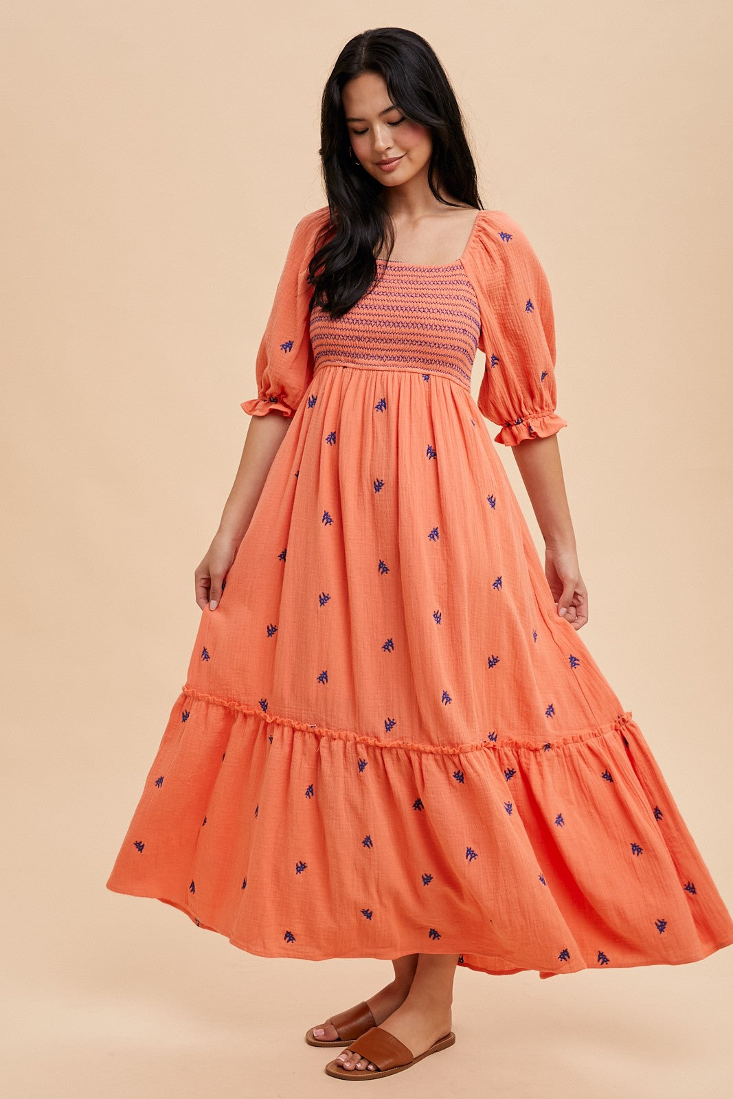 woman wearing an orange maxi dress with puff sleeves, smocked bodice, and floral embroidery, standing against a light backdrop