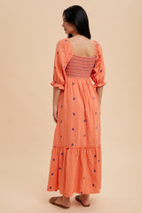 woman wearing an orange maxi dress with puff sleeves and floral embroidery, showing a smocked back and tiered hemline