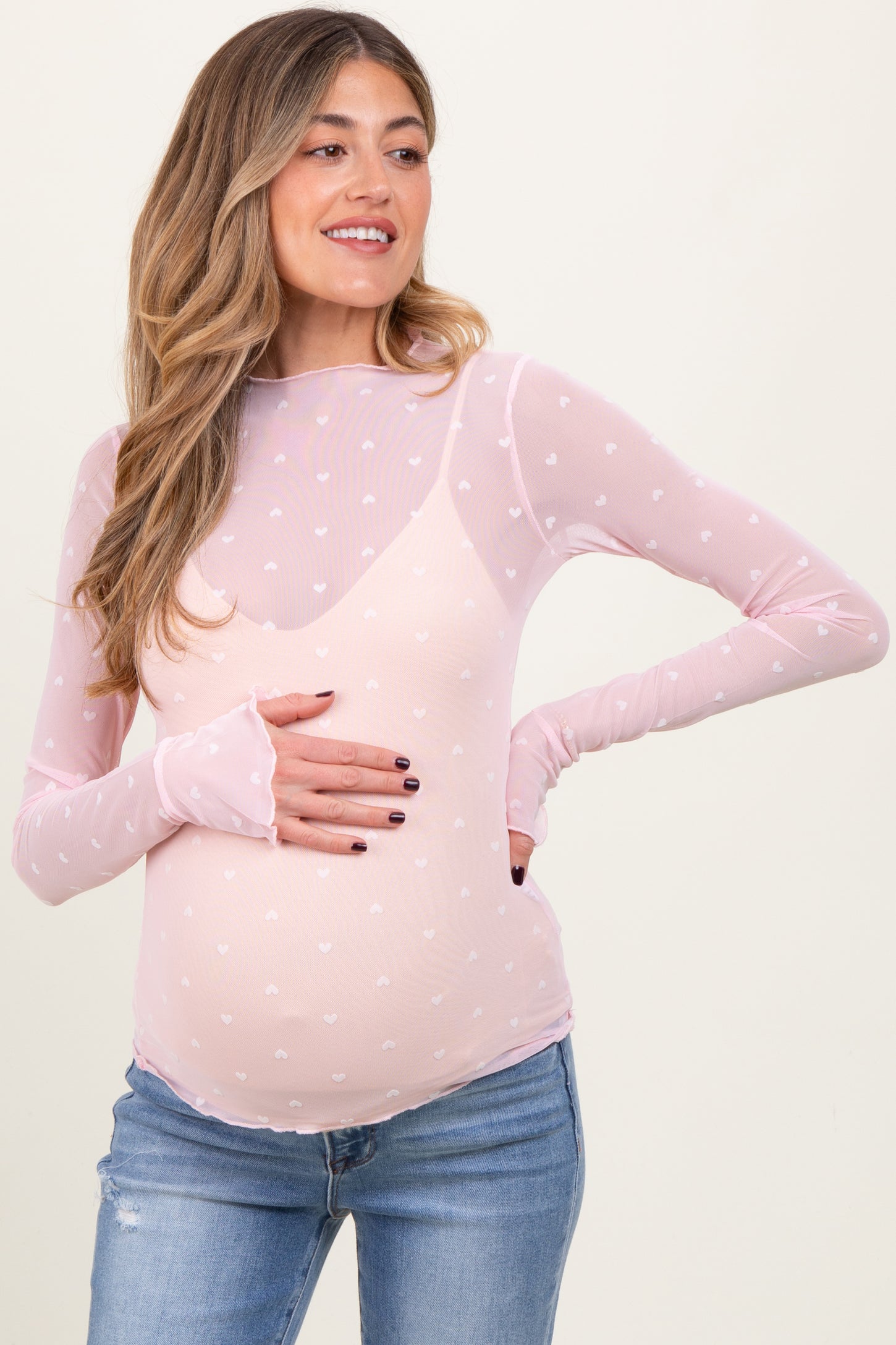 woman wearing a light pink long sleeve maternity top with white heart patterns and gently holding her baby bump