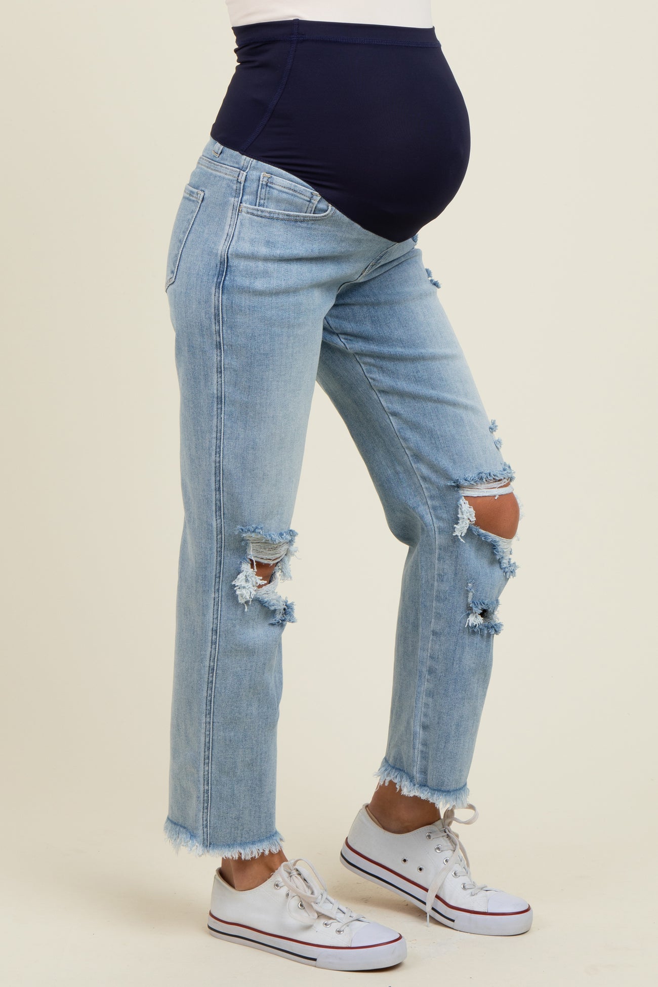 woman wearing light blue distressed maternity jeans with a navy belly panel and sneakers, showing her baby bump