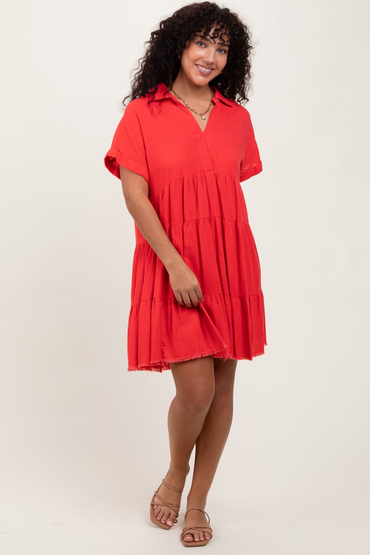 woman wearing a red tiered short sleeve dress with a collar and frayed hem, standing with one hand on her dress