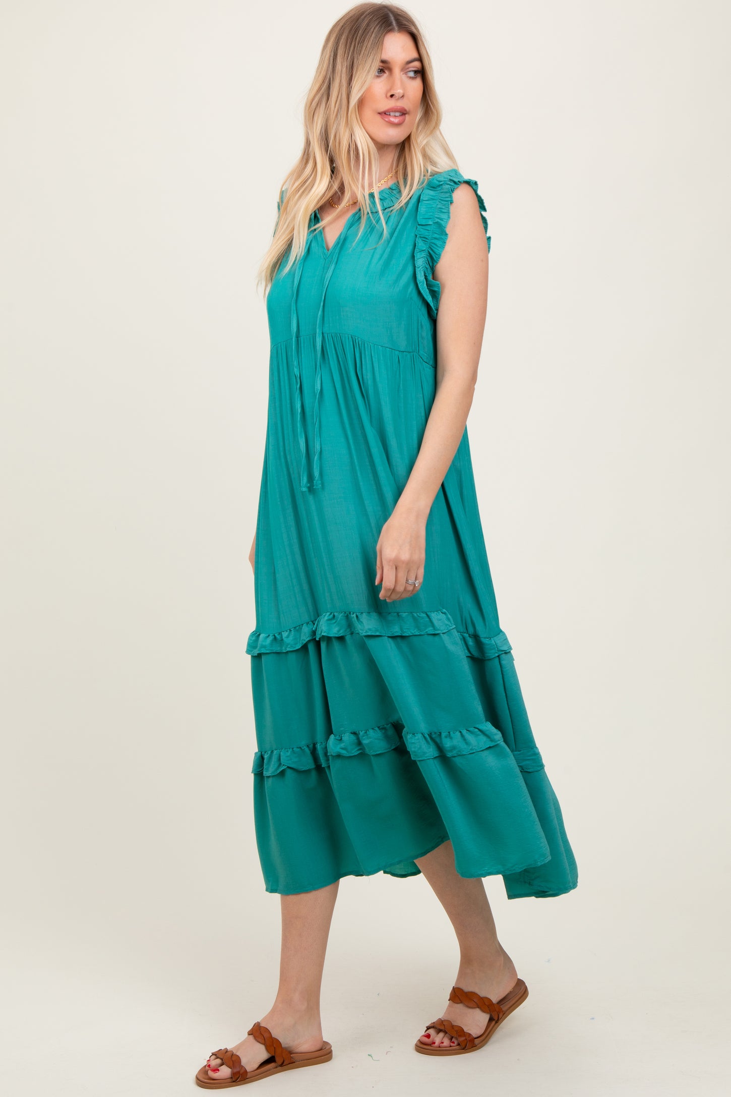 woman wearing a teal maxi dress with ruffle details and tie neck, standing against a plain background
