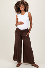 woman wearing a white sleeveless maternity top and brown wide-leg pants, gently cradling her baby bump