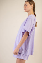 woman wearing a lilac oversized t-shirt with side slits and matching shorts, standing sideways against a neutral background