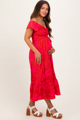 woman wearing a bright pink floral maternity dress with off-shoulder sleeves and a ruffled hem, holding her baby bump