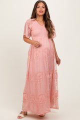 woman wearing a light pink maternity maxi dress with floral embroidery and short flutter sleeves, showcasing her baby bump