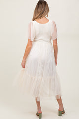 woman wearing a white maternity midi dress with sheer puff sleeves and a tiered tulle skirt, showing her baby bump