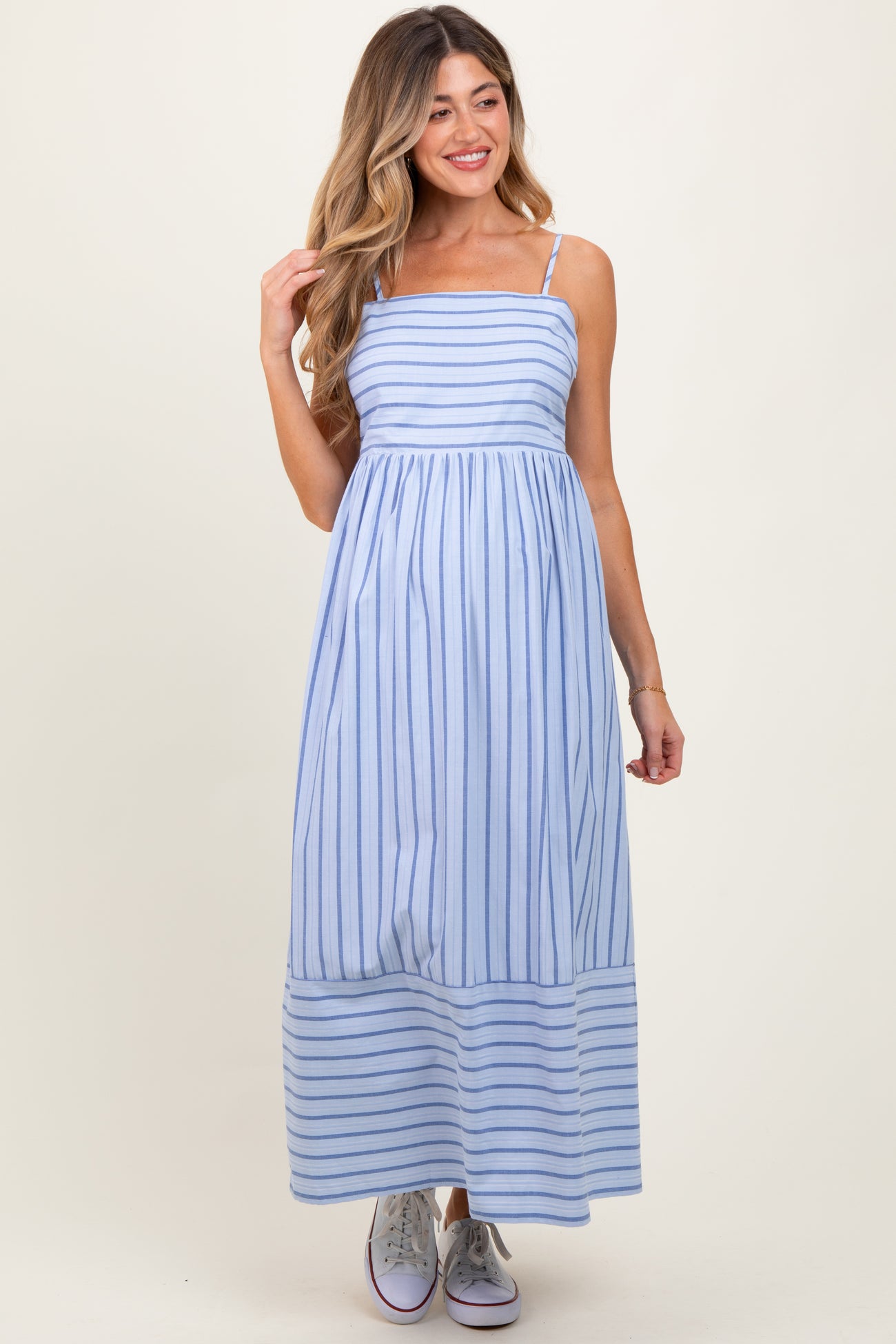 woman wearing a light blue striped maternity maxi dress with thin straps and a flowing skirt, holding her hair