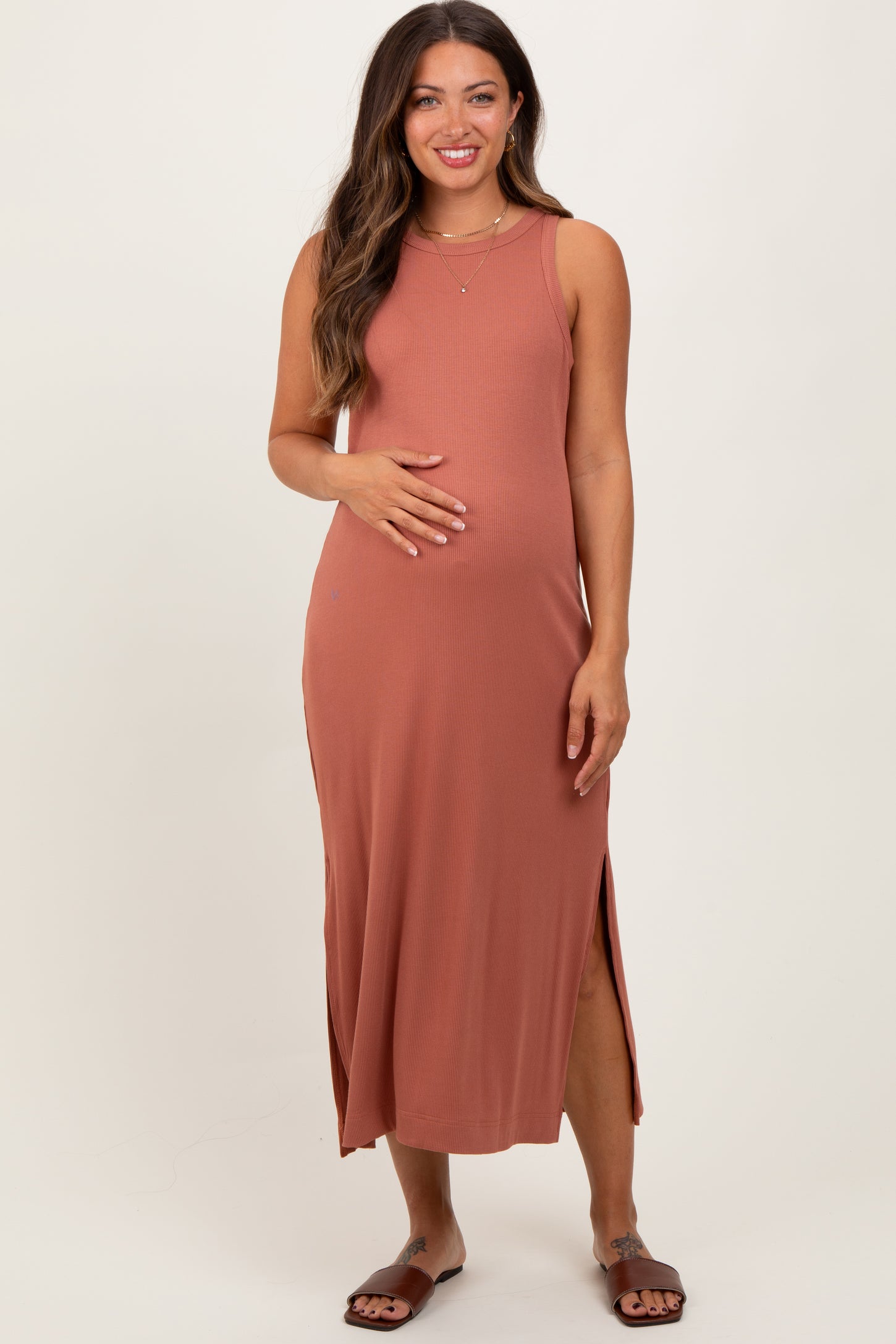 woman wearing a rust-colored sleeveless maternity maxi dress with side slits, gently cradling her baby bump