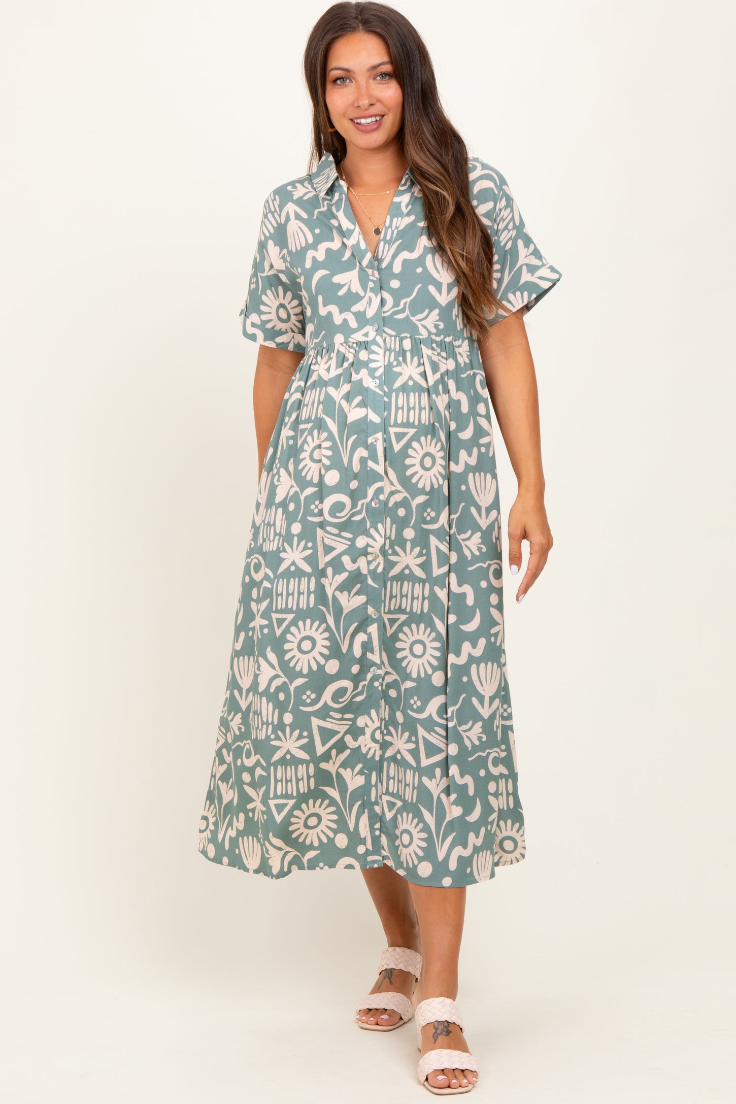 woman wearing a green floral print maternity dress with short sleeves and a button-down front, smiling while standing