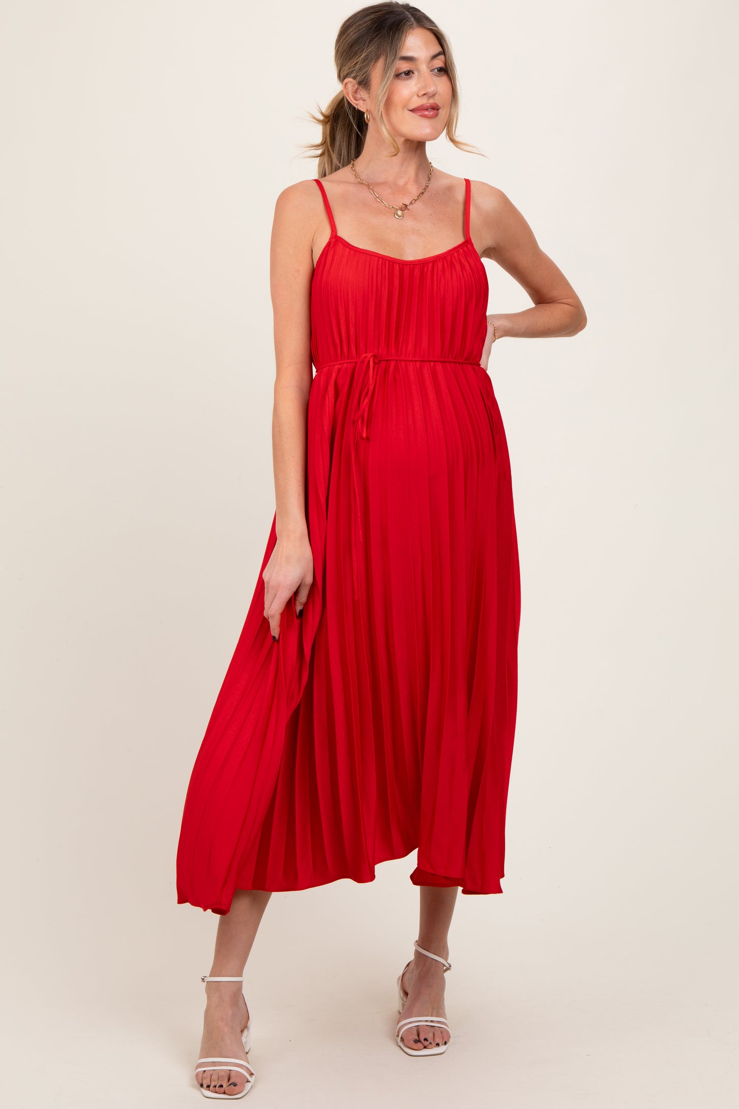 woman wearing a red pleated maternity dress with adjustable straps and a tie at the waist, showcasing her baby bump