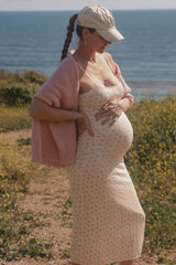 woman wearing a sleeveless floral maternity dress and a pink cardigan, showing her baby bump by the ocean
