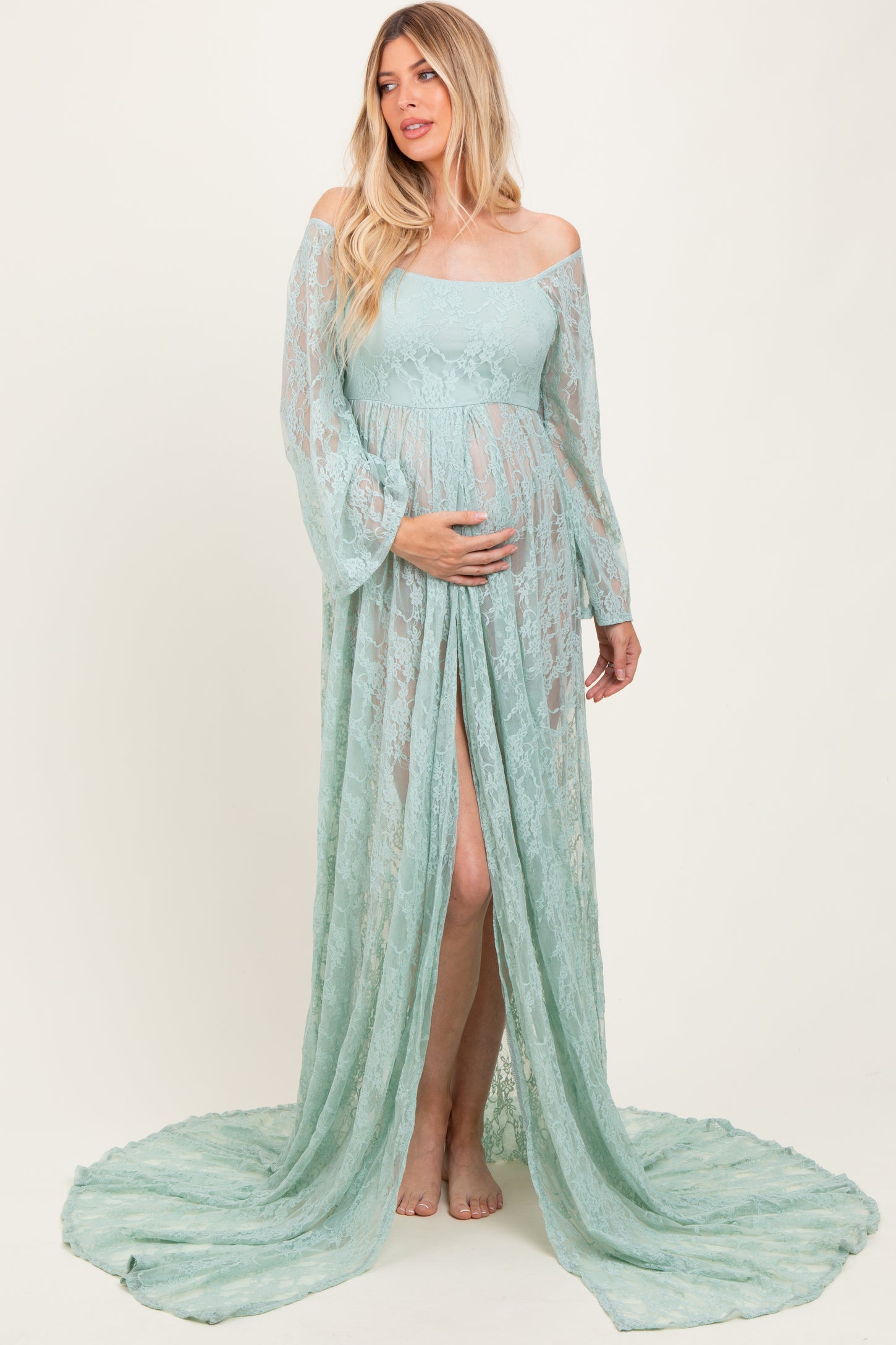 woman wearing a light green lace maternity gown with off-the-shoulder sleeves and a side slit, holding her baby bump