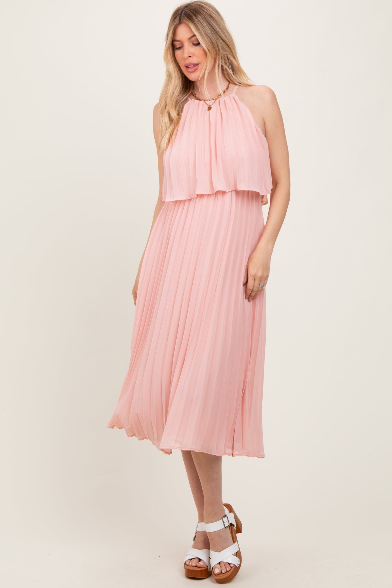 woman wearing a light pink pleated midi dress with a high neckline and layered bodice, styled with white sandals
