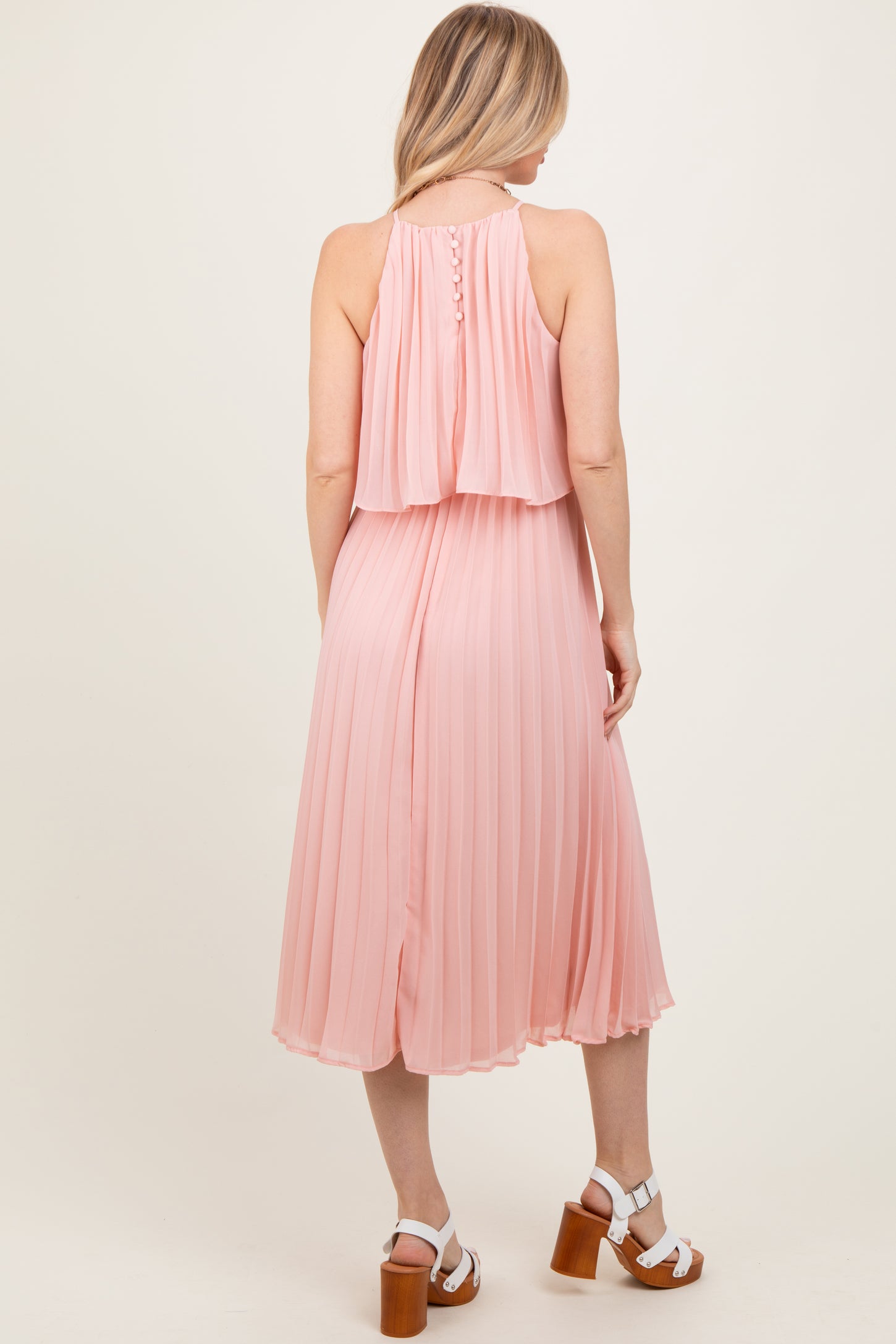 woman wearing a blush pink pleated midi dress with a halter neck and layered back, paired with white heeled sandals