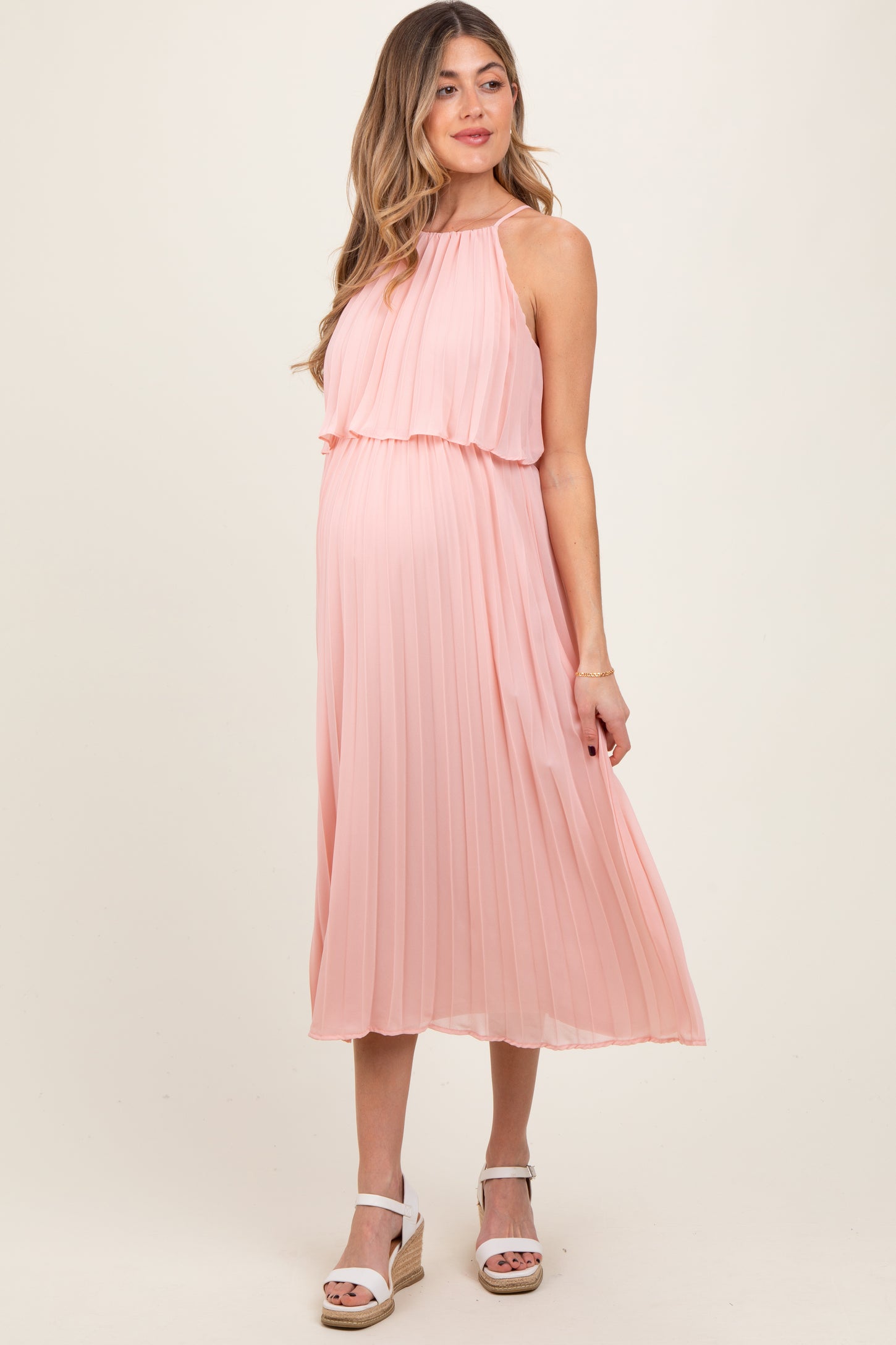 woman wearing a blush pink pleated maternity dress with a flowing halter neckline and visible baby bump