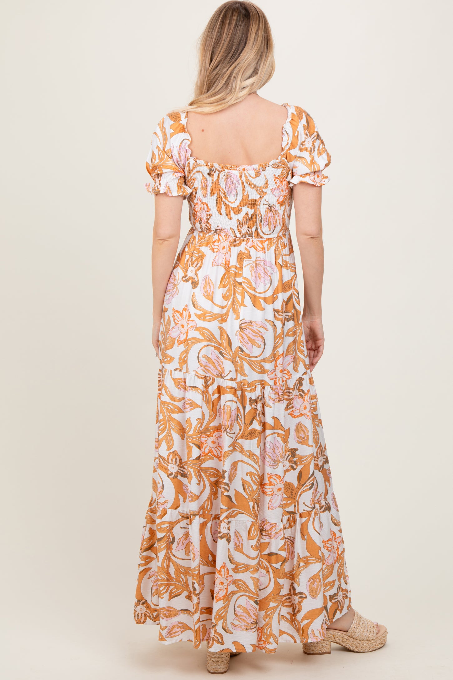 woman wearing a tan and pink floral print maxi dress with short puff sleeves and a smocked bodice, viewed from the back