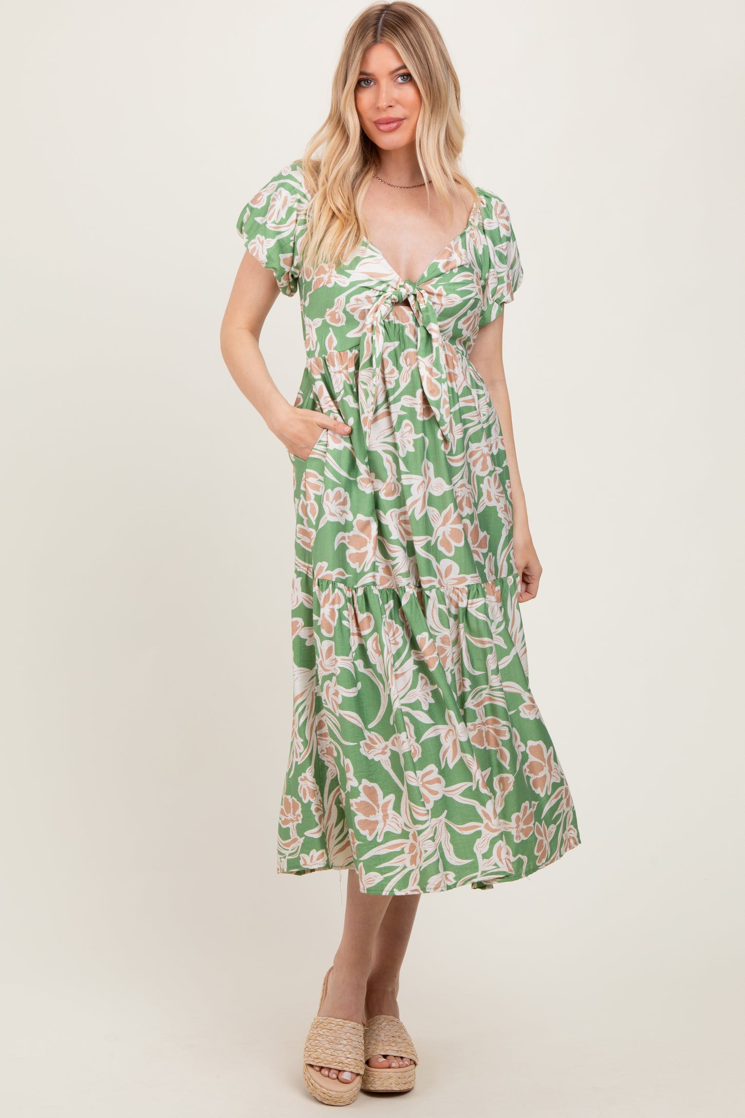 woman wearing a green floral print maxi dress with a tie front and short sleeves, standing and posing with hands in pockets