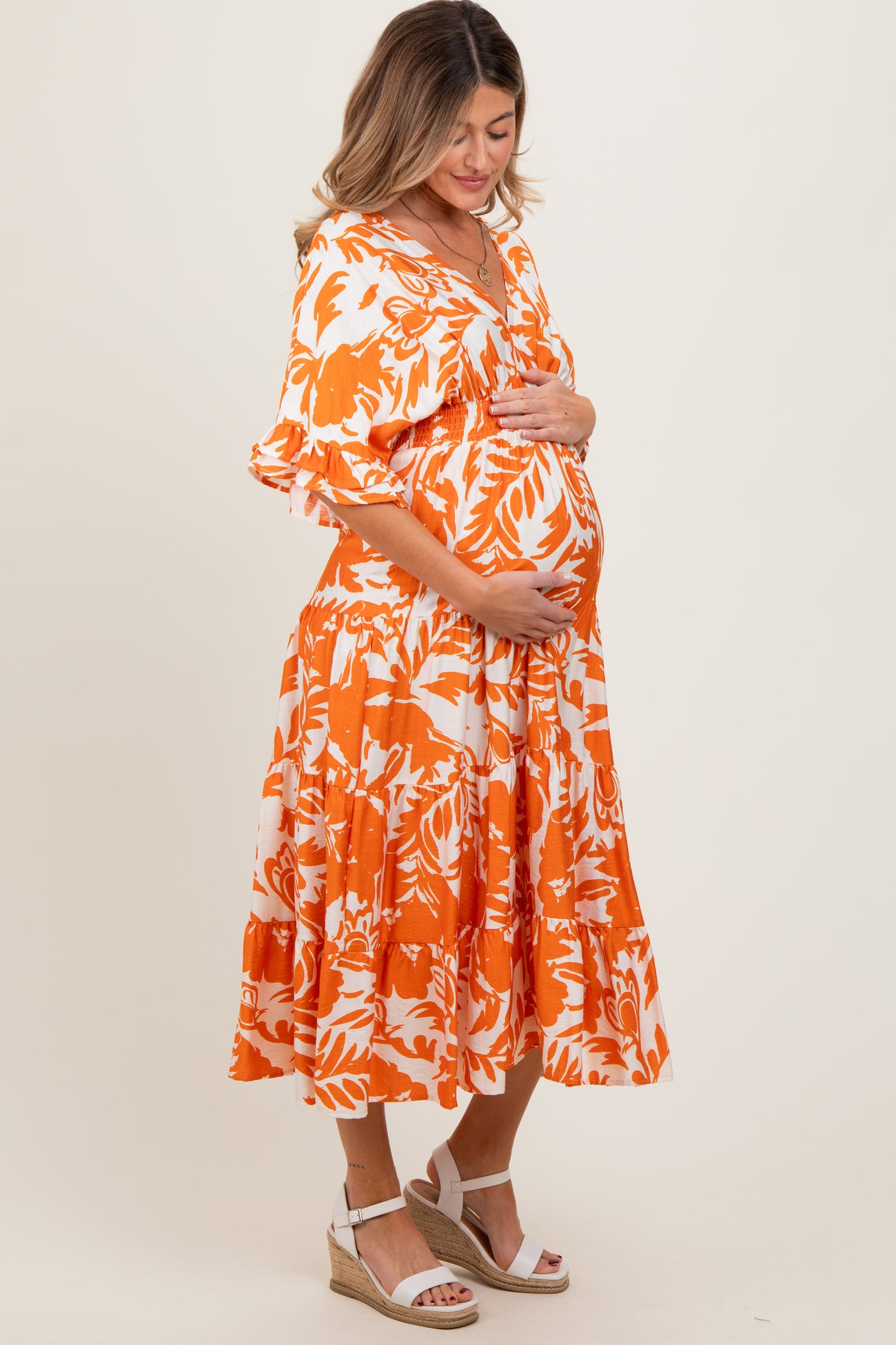 woman wearing an orange and white floral maternity dress and cradling her baby bump