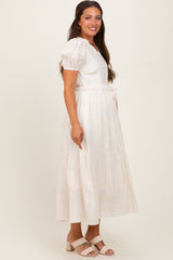 woman wearing a cream maternity midi dress with puff sleeves and a tie waist, featuring a tiered skirt and button details