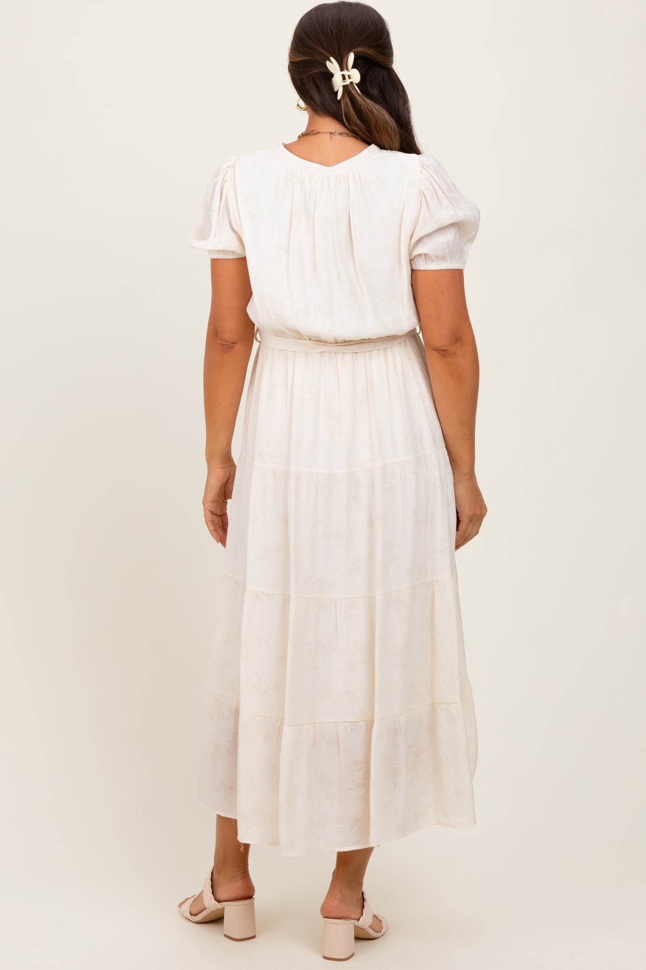 woman wearing a light cream maternity dress with puff sleeves and a tiered skirt, showing her baby bump from the back