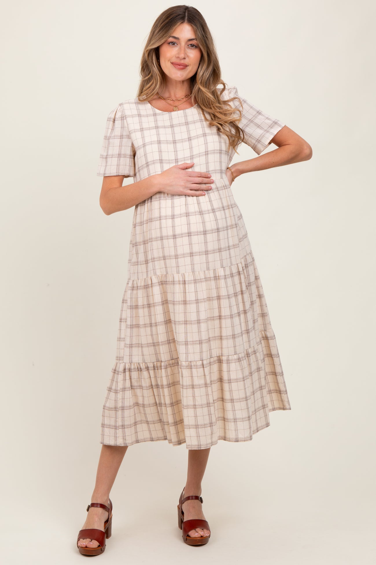 woman wearing a beige and brown plaid maternity dress with tiered hem and holding her baby bump