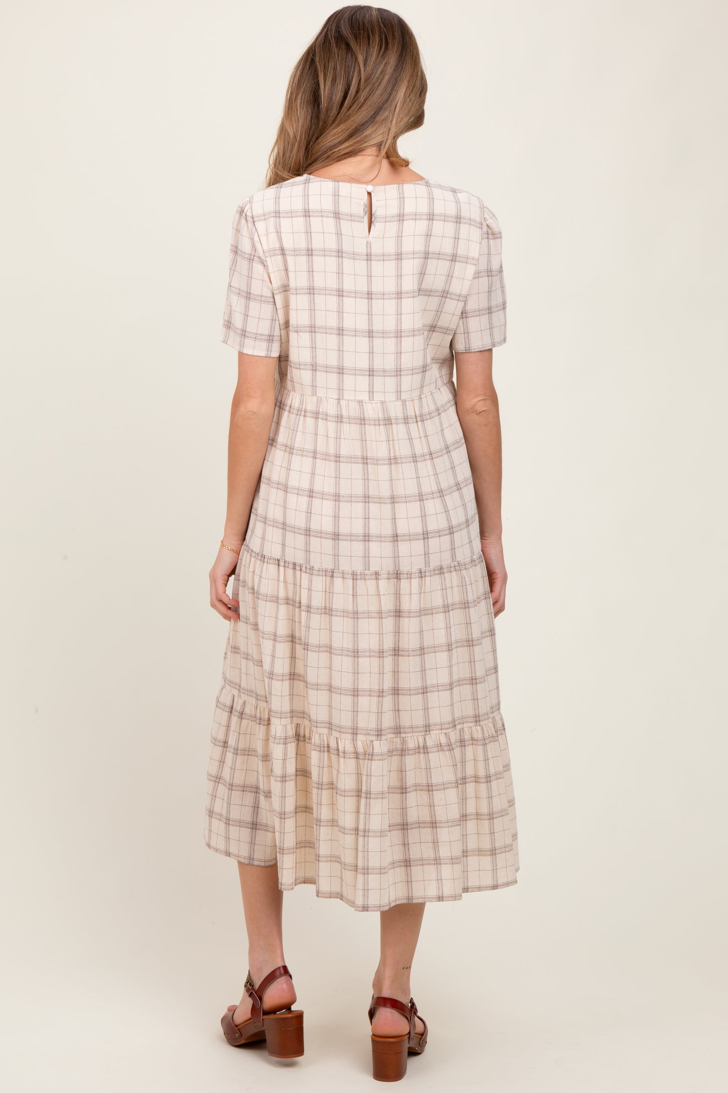 woman wearing a beige and brown plaid maternity dress with short sleeves and tiered skirt, standing with back to the camera