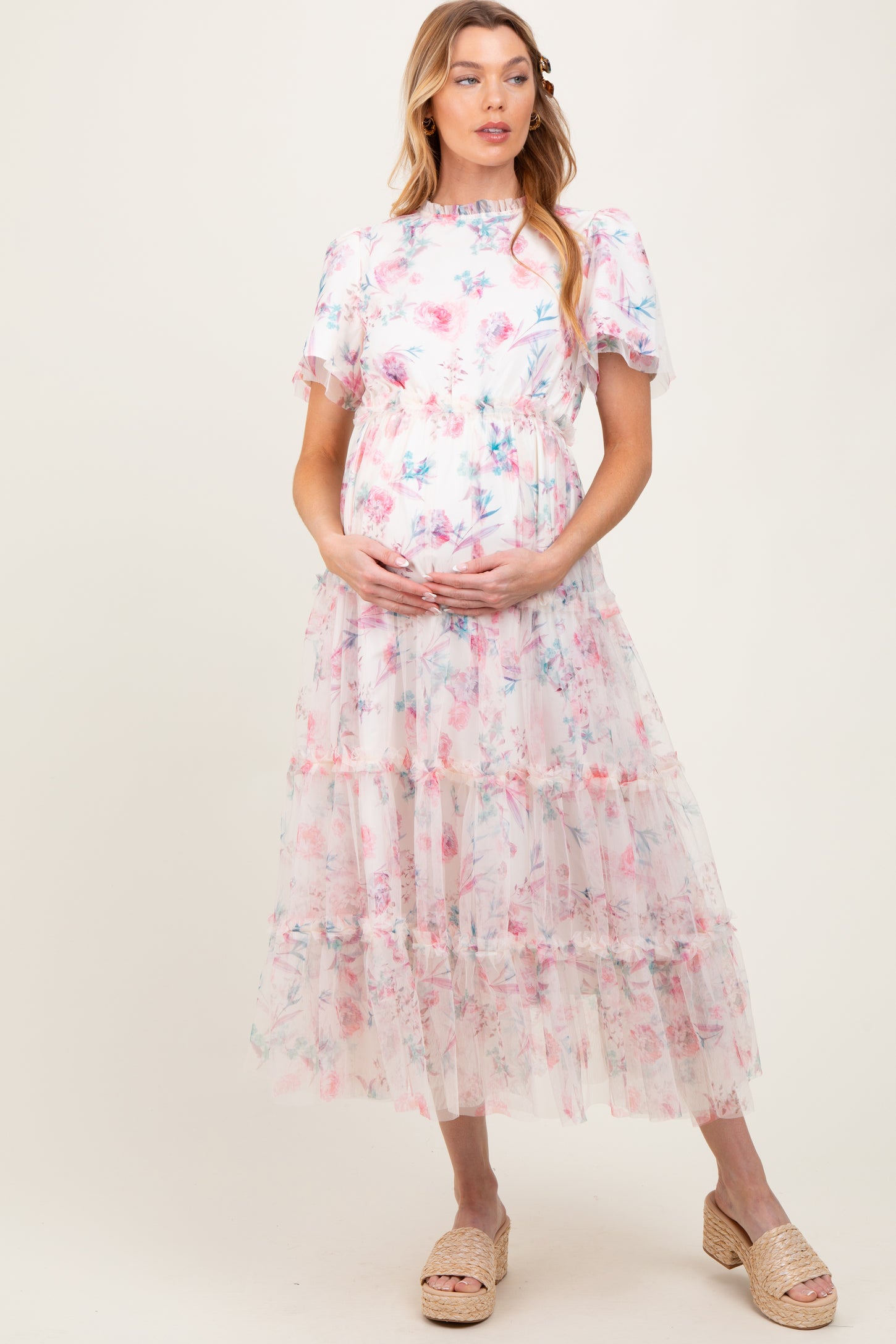 woman wearing a floral print pastel pink maternity dress with ruffled sleeves and tiers, holding her baby bump