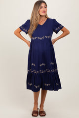 woman wearing a navy blue maternity dress with embroidered floral details and a tiered hem, styled with sandals