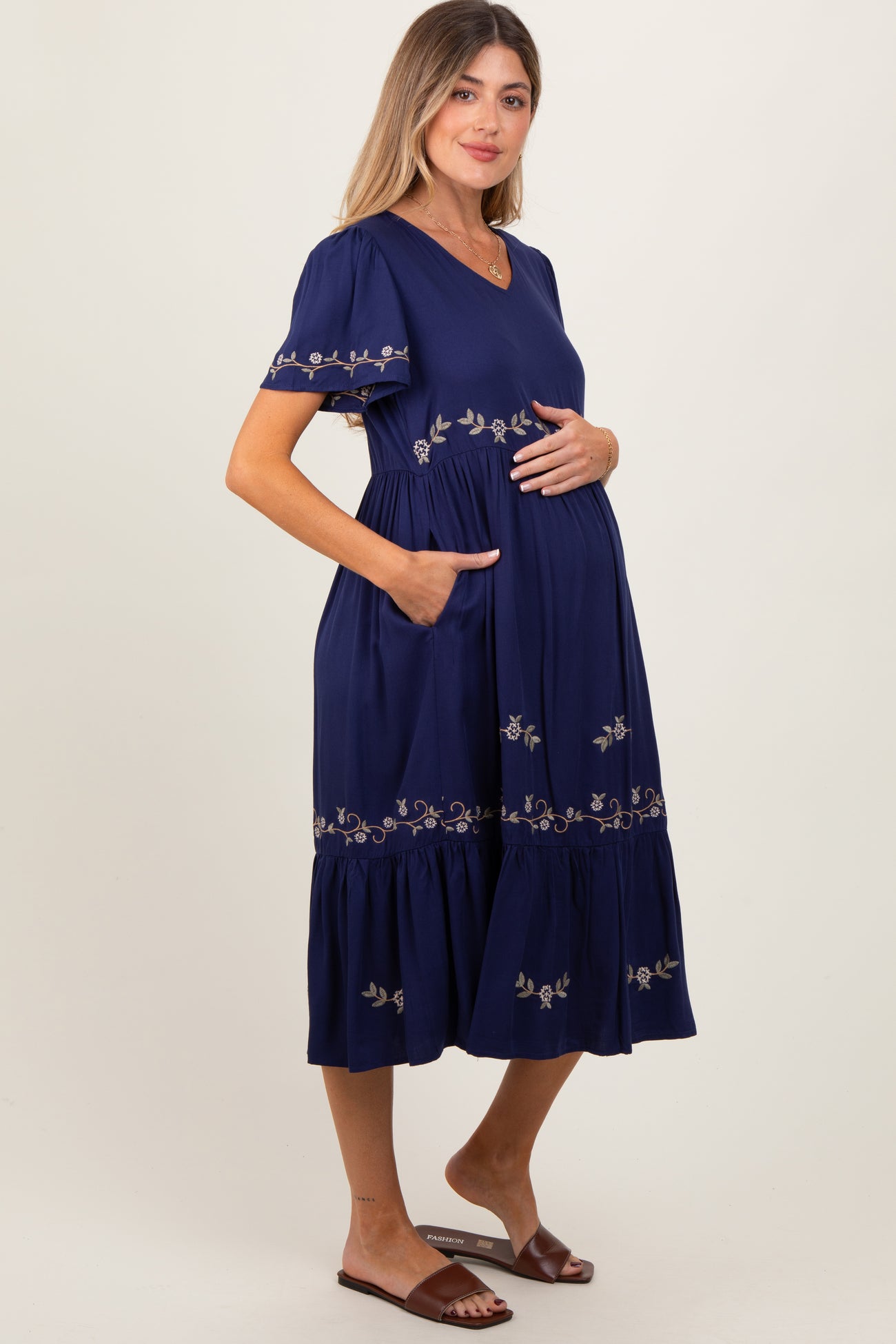 woman wearing a navy blue maternity dress with floral embroidery and pockets, cradling her baby bump