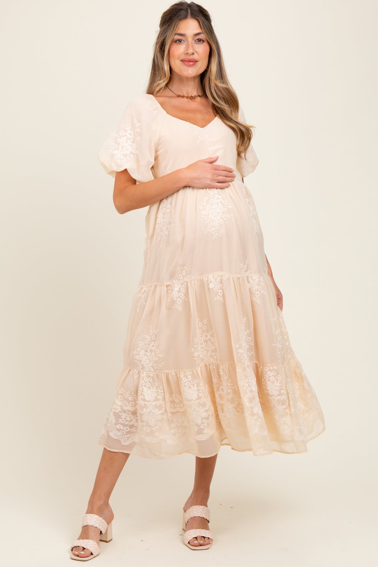 woman wearing a beige lace embroidered maternity dress with short sleeves and a tiered hem, holding her baby bump