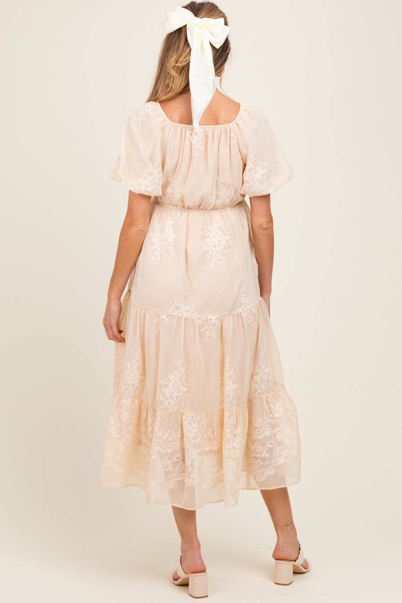 woman wearing a peach-colored maternity dress with floral lace details and a tiered skirt, seen from the back