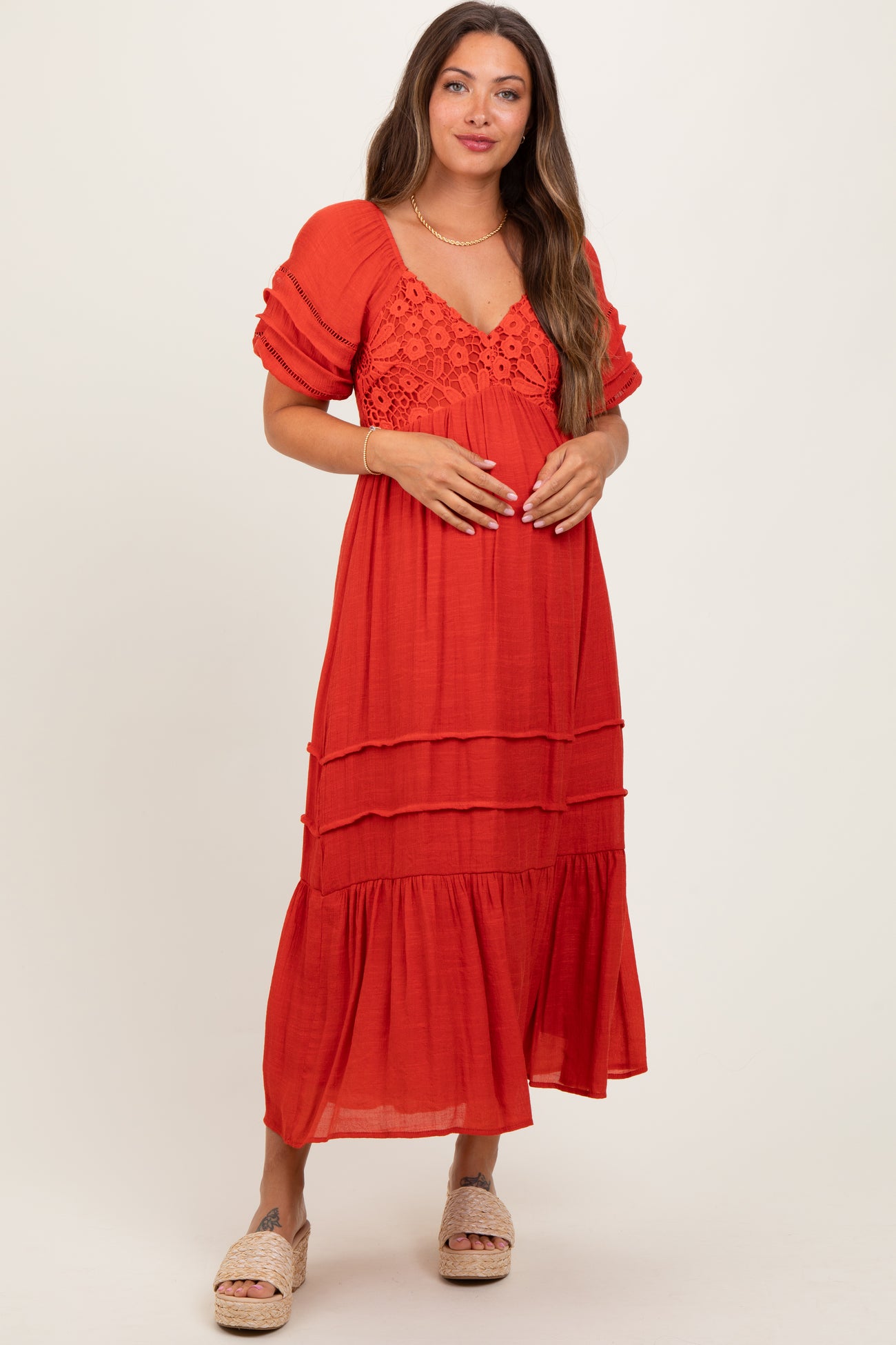woman wearing a red tiered maternity dress with puff sleeves and lace detailing, gently holding her baby bump