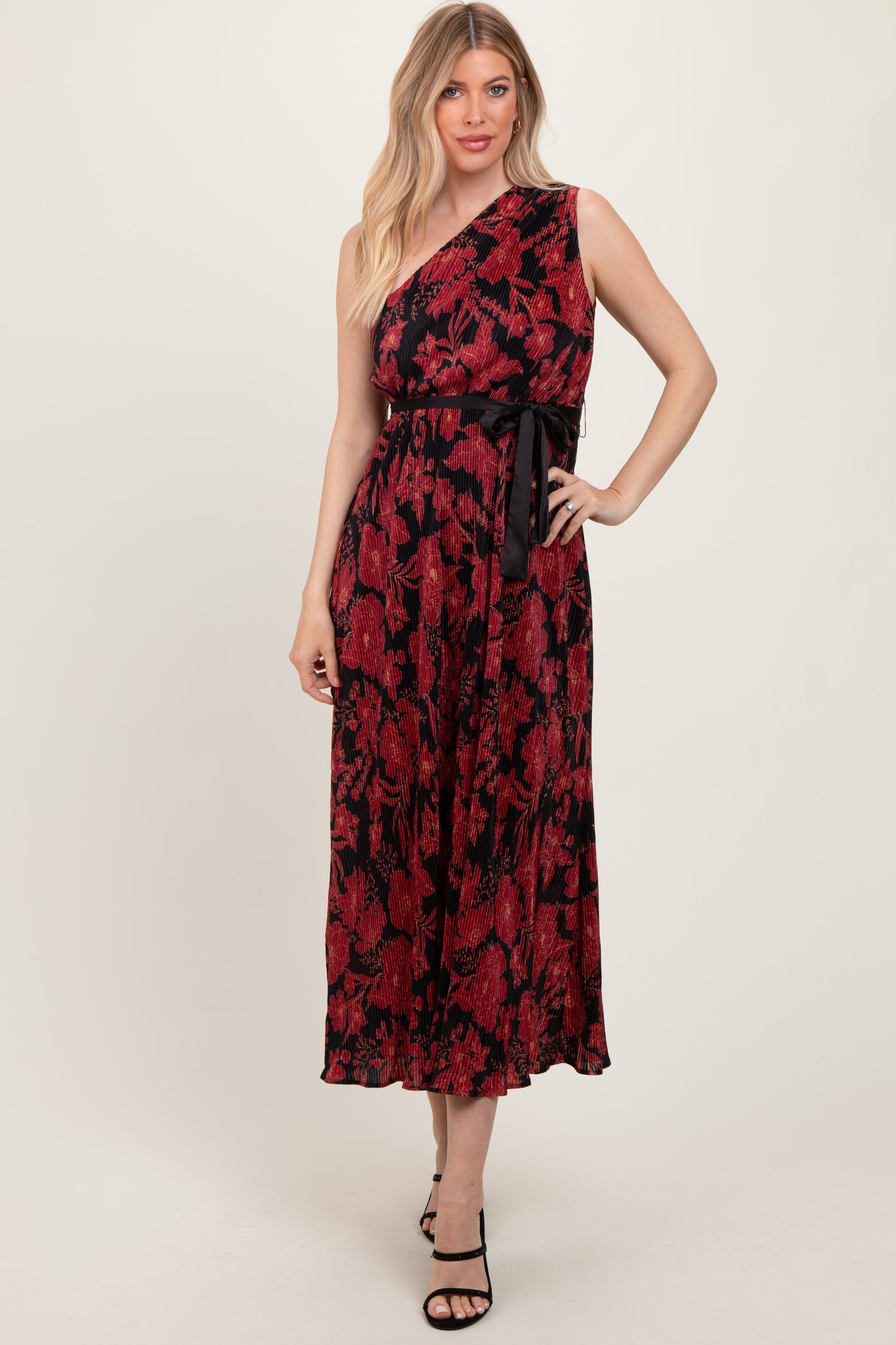 woman wearing a red and black floral one-shoulder maxi dress with a tied waist detail and strappy black heels