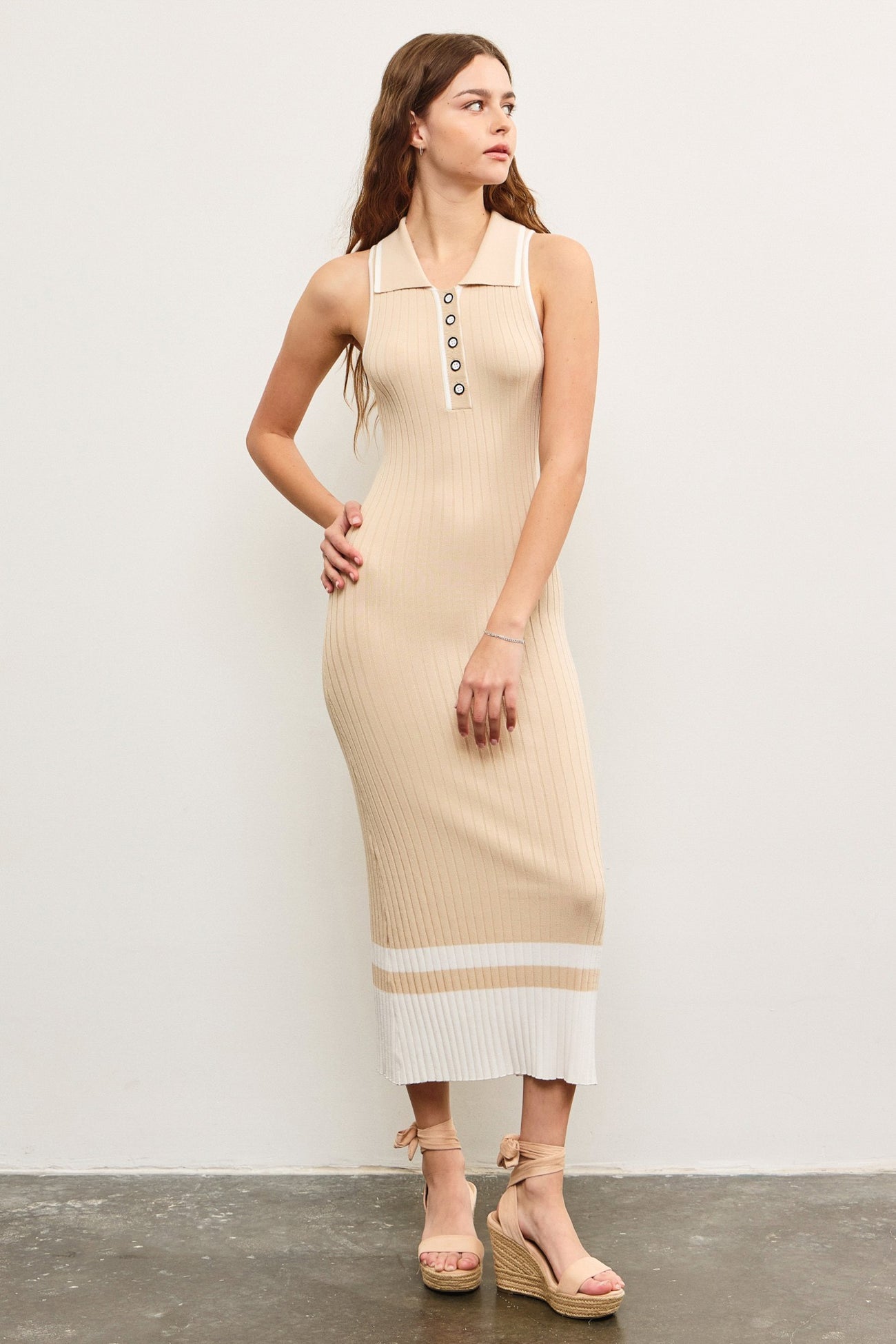woman wearing a beige ribbed knit sleeveless midi dress with a polo collar and button details, standing against a white wall