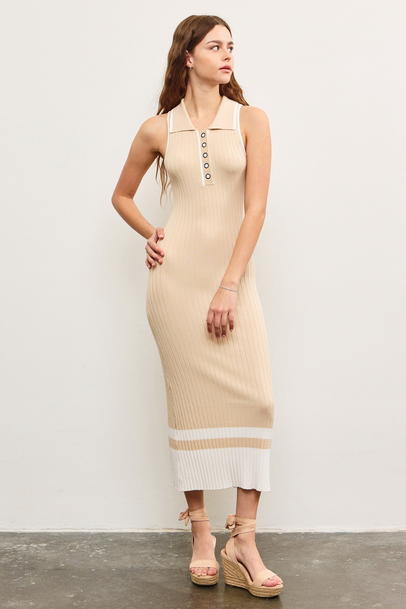 woman wearing a beige ribbed knit sleeveless midi dress with a polo collar and button details, standing against a white wall