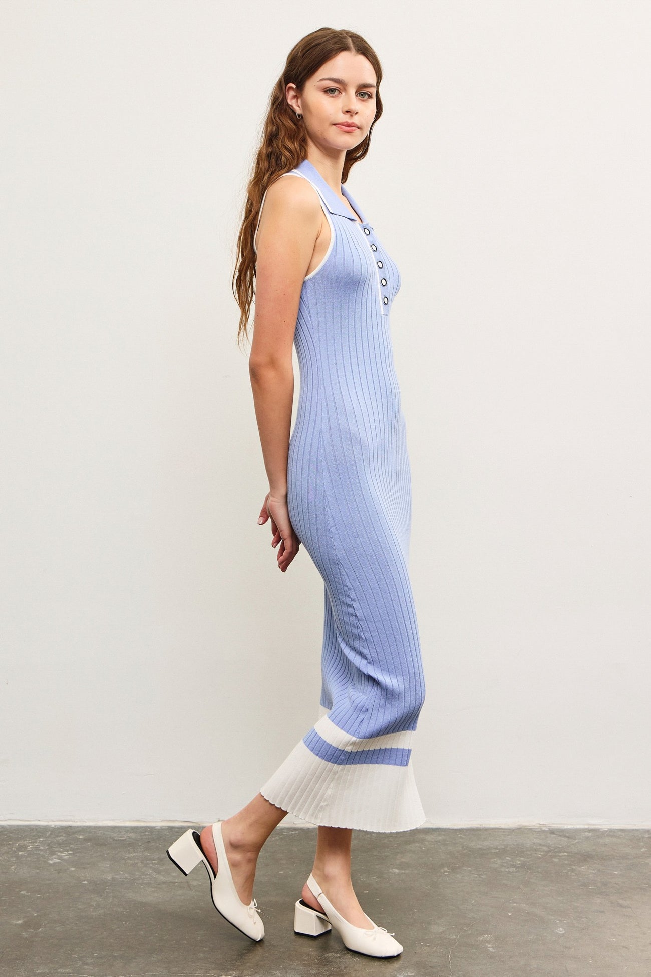woman wearing a light blue ribbed knit midi dress with buttons and a white hem, standing sideways on a gray floor