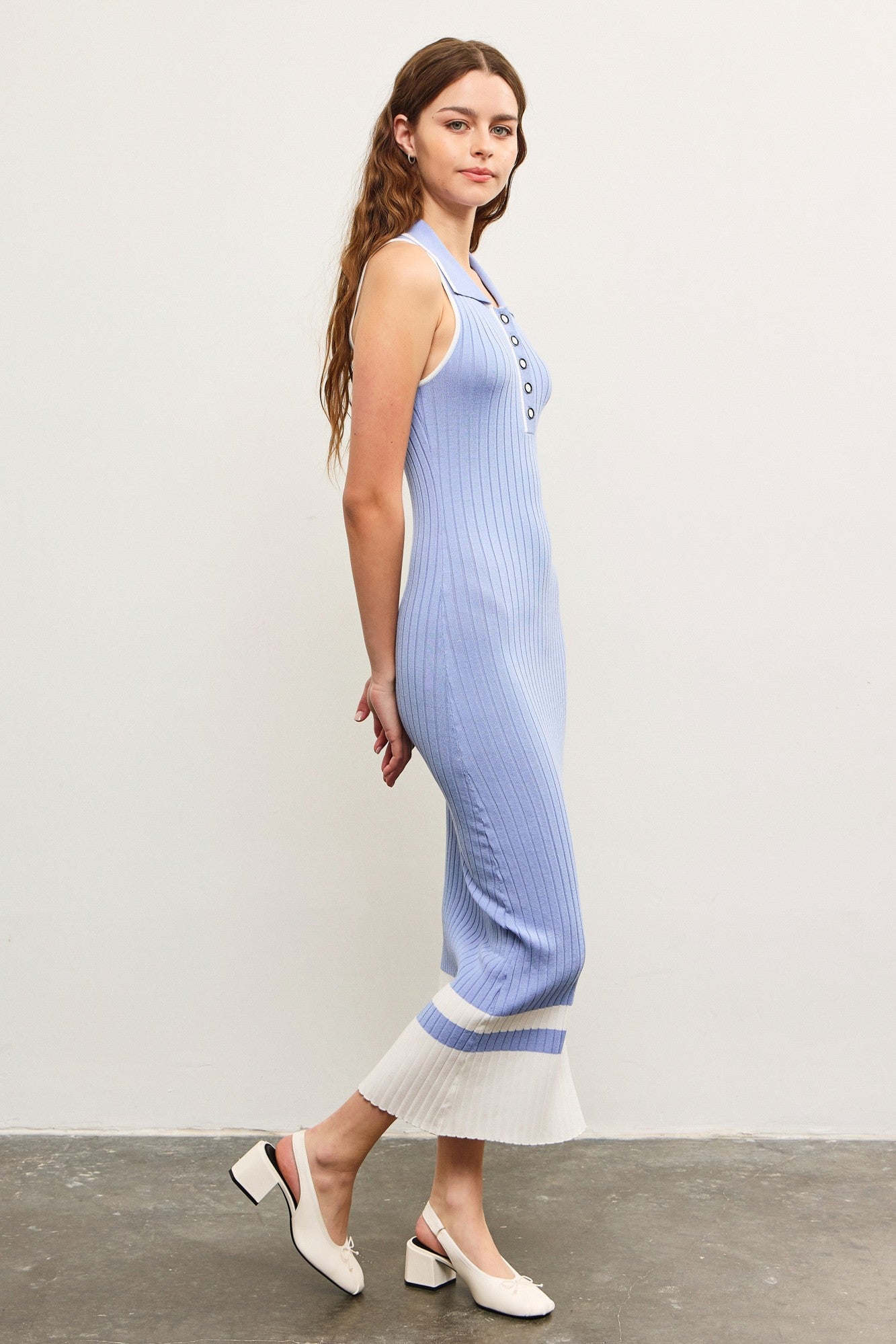 woman wearing a light blue ribbed knit midi dress with buttons and a white hem, standing sideways on a gray floor