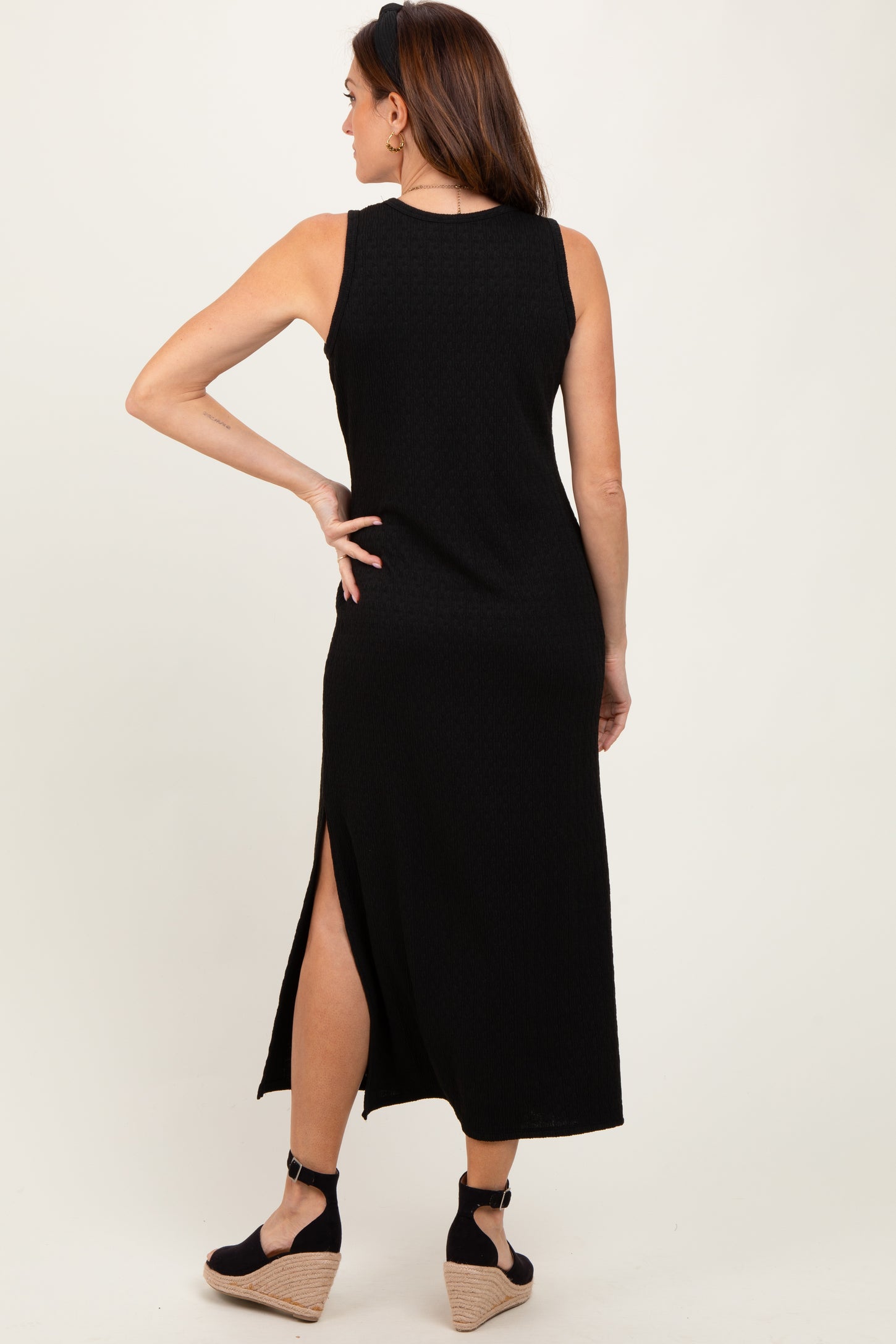 woman wearing a black sleeveless maxi dress with a side slit, showing the back view and a pair of wedges