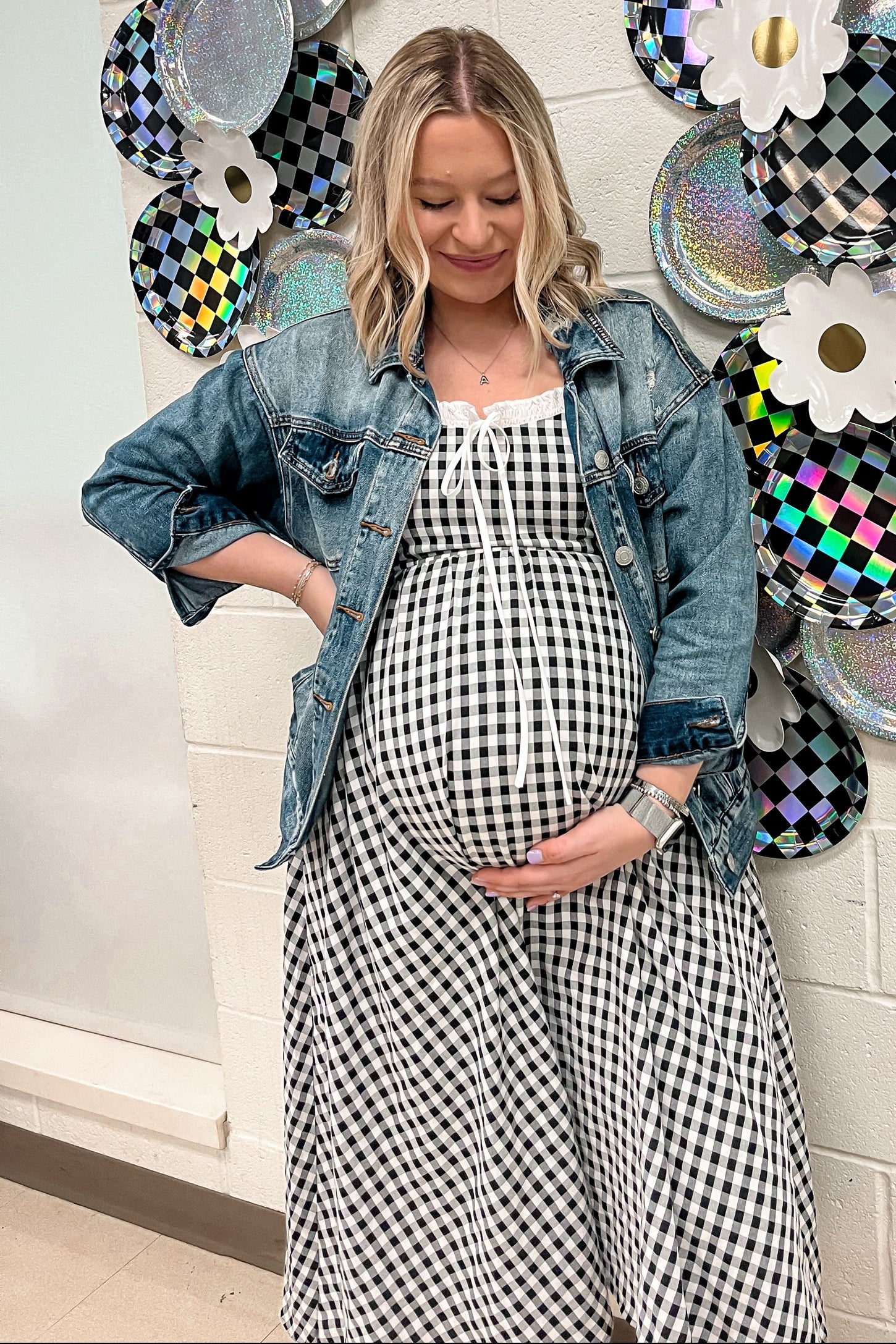 woman wearing a black and white gingham maternity dress with a denim jacket and holding her baby bump