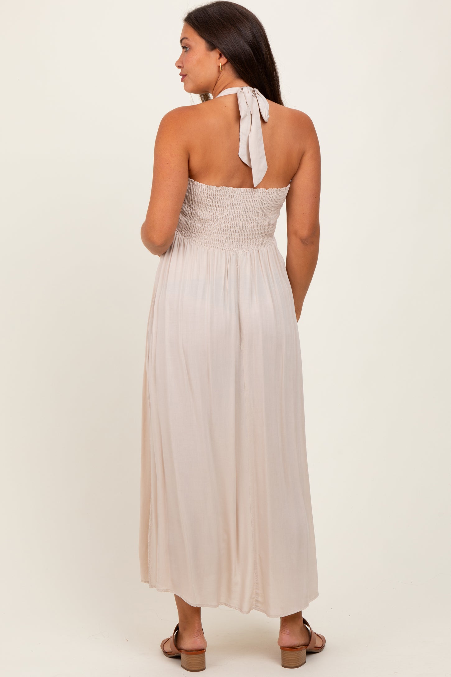 woman wearing a light beige maternity maxi dress with a smocked bodice and halter tie detail, back view