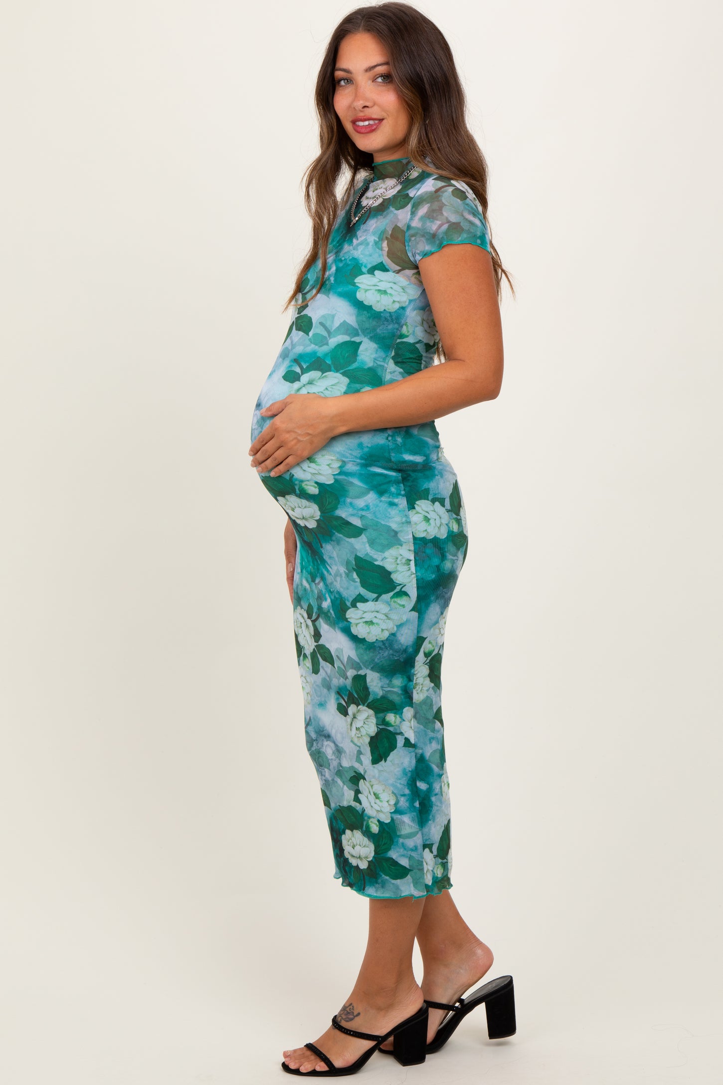 woman wearing a floral green maternity dress, hugging her baby bump, standing in side profile with a smile