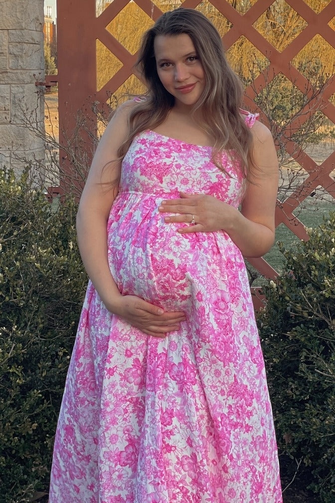 woman wearing a pink floral maternity dress with a visible baby bump, holding her belly outdoors