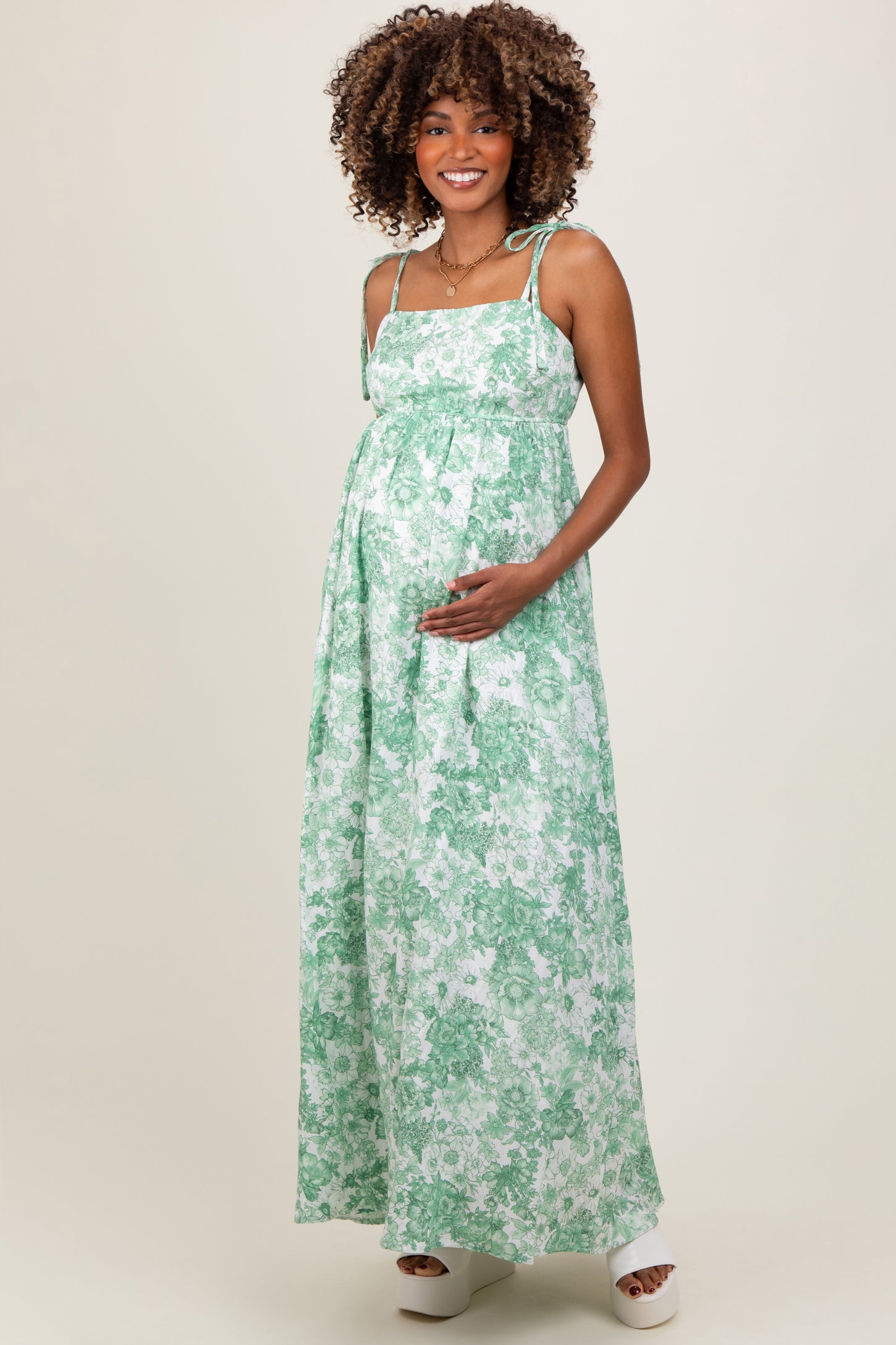 woman wearing a green floral maternity maxi dress, showing her baby bump and smiling with hands on her belly