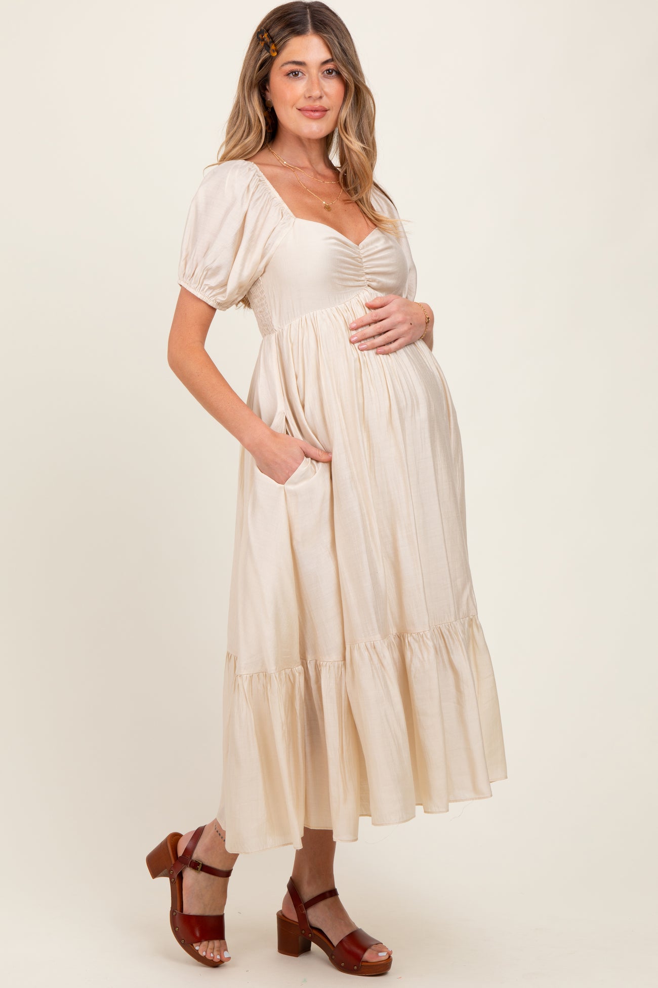 woman wearing a beige maternity dress with puff sleeves and front ruching, holding her baby bump and standing in a relaxed pose