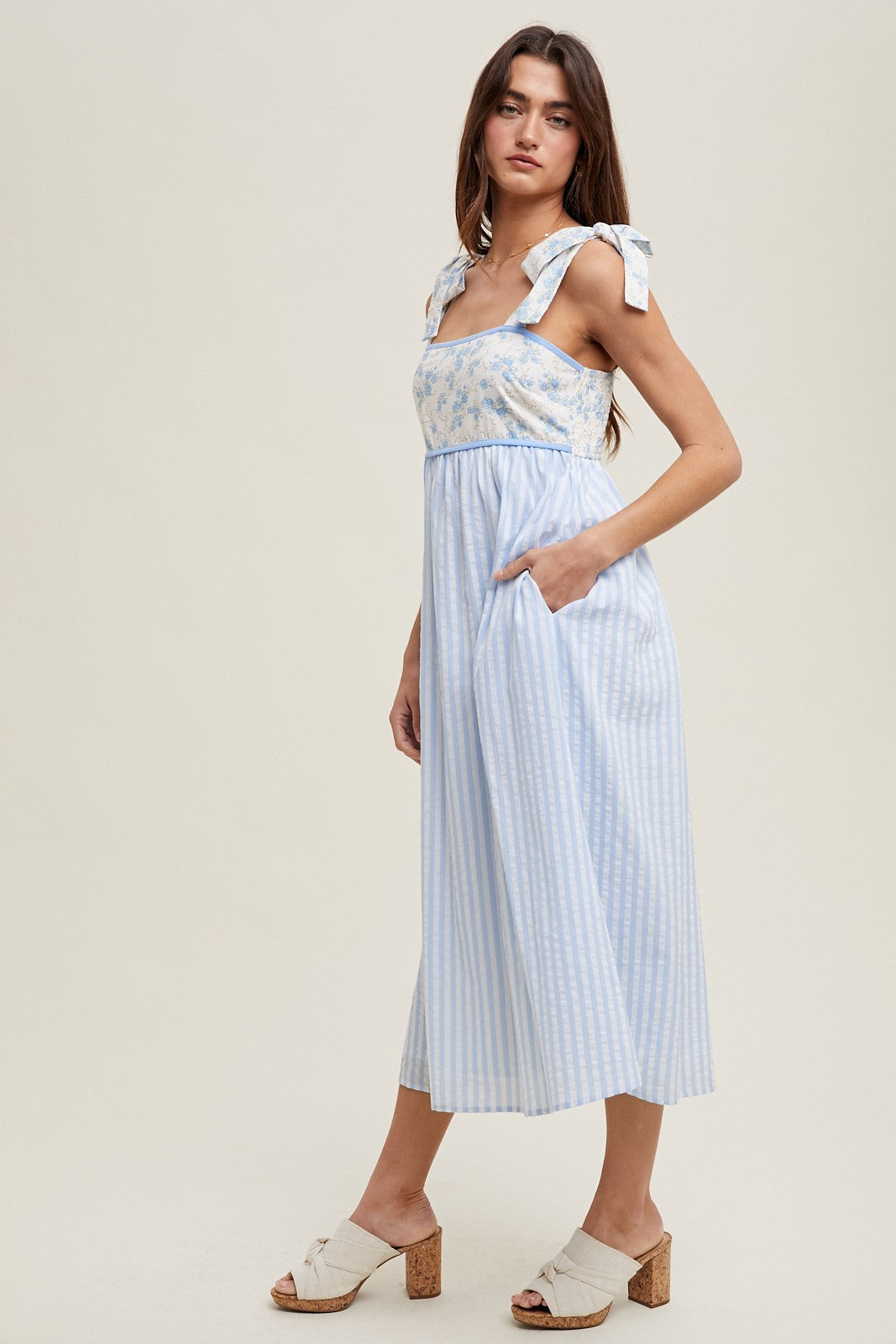 woman wearing a light blue sleeveless maxi dress with floral and striped patterns and tie shoulder details