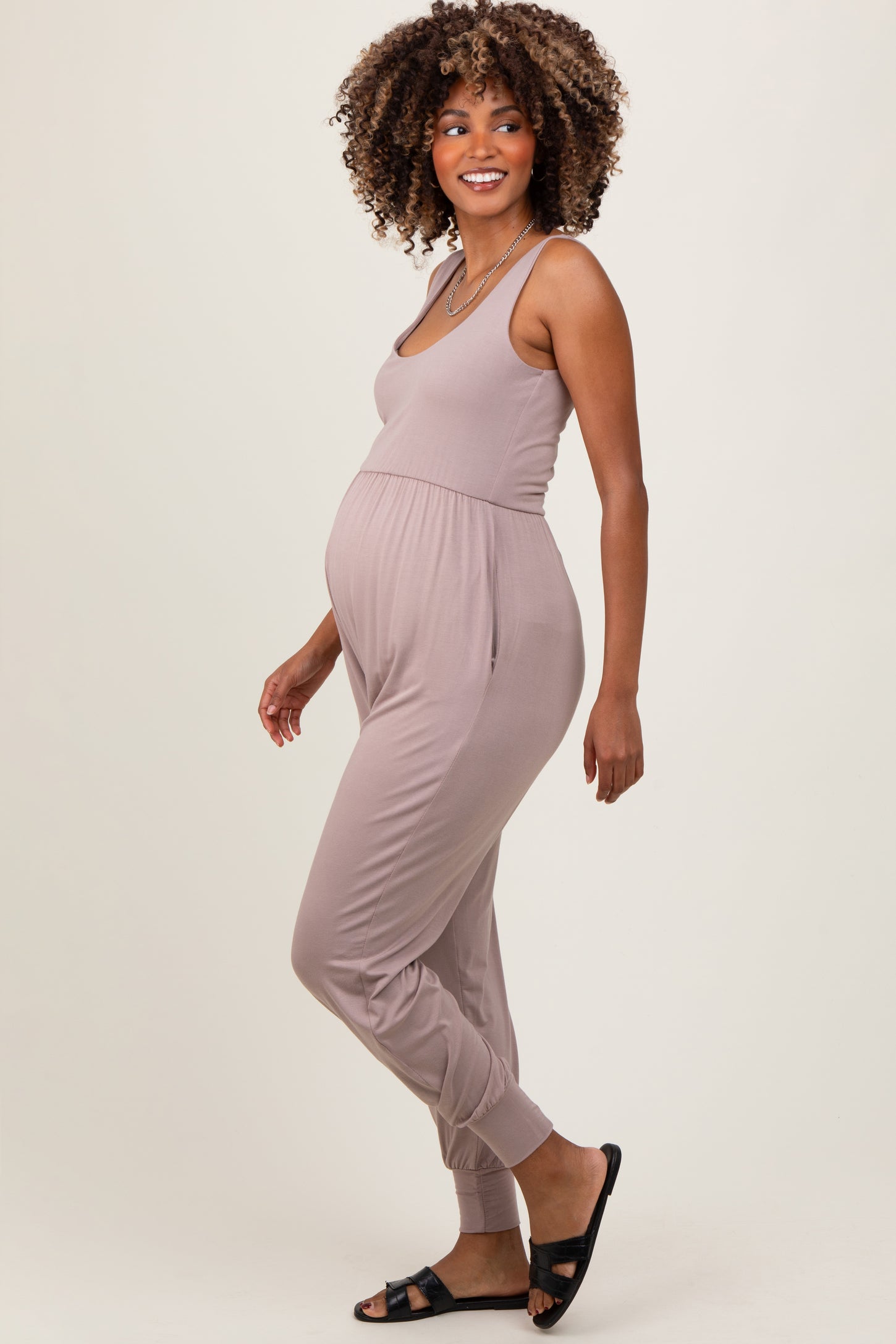 woman wearing a light taupe maternity jumpsuit with a fitted design and elastic ankle cuffs, showing her baby bump