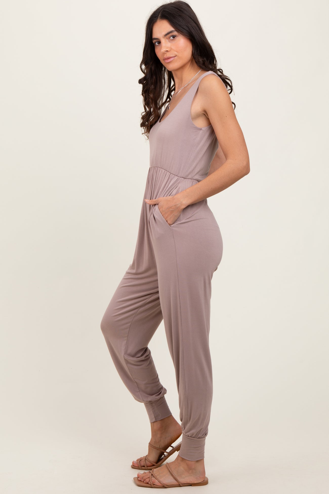 woman wearing a taupe sleeveless jumpsuit with pockets and fitted cuffs, standing sideways